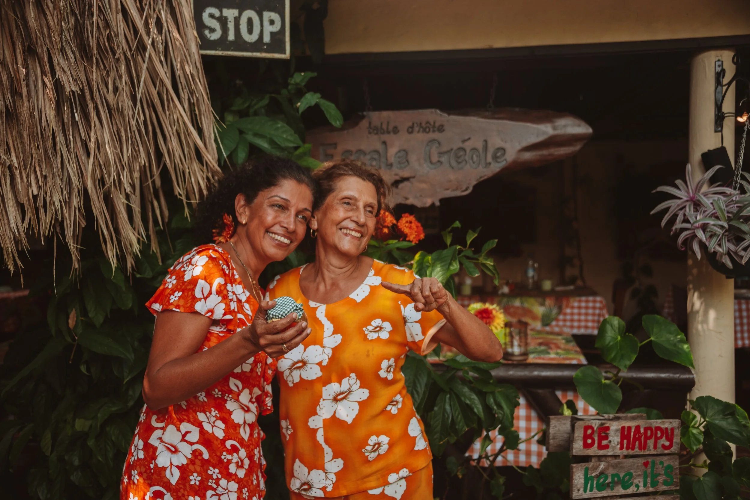 Two women standing outdoors, smiling and posing for a photo at a tropical-themed location with lush green plants and colorful flowers. One woman is holding a small item, and both are dressed in bright, floral-patterned dresses.