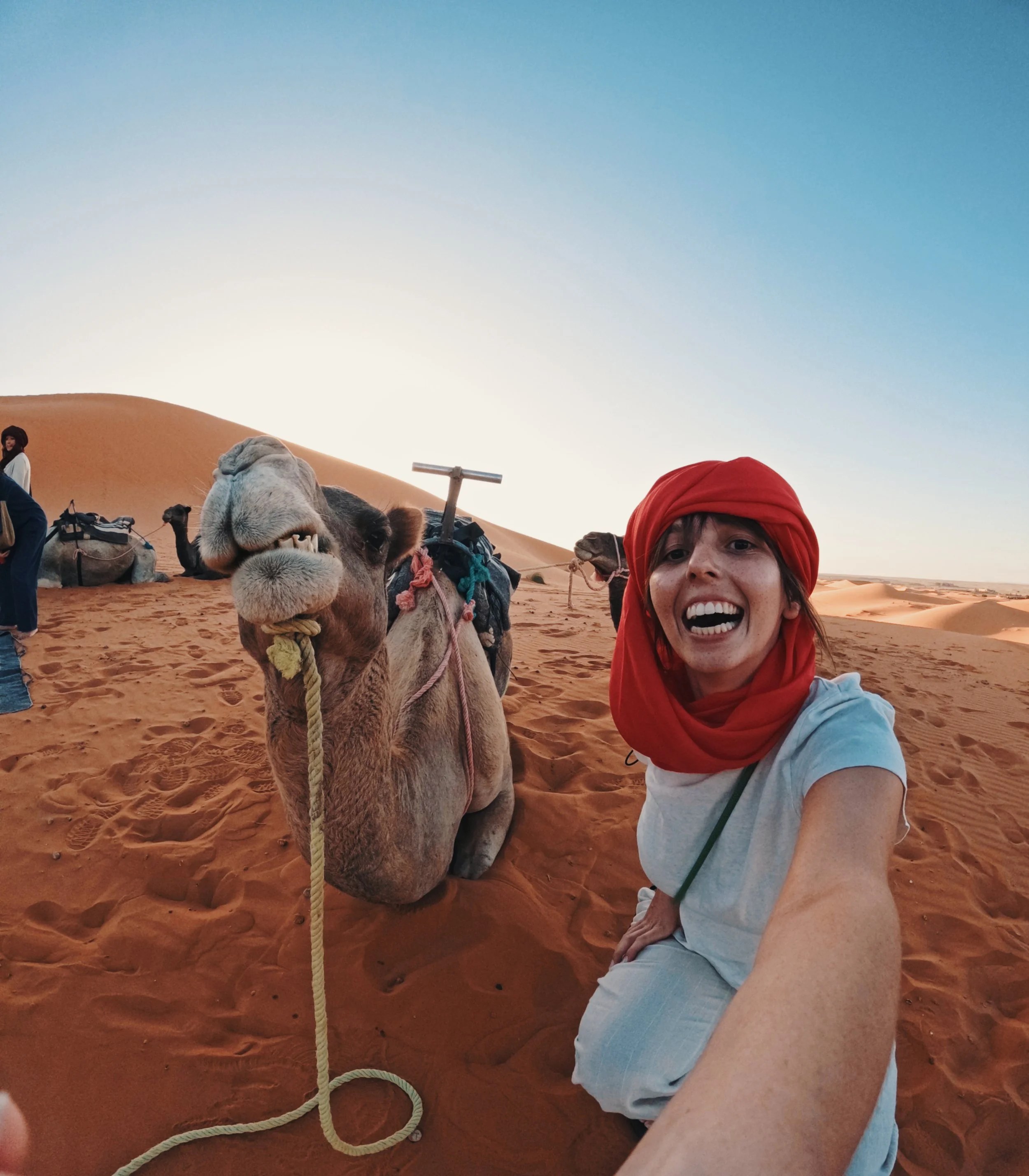A woman taking a selfie with a camel in a desert at sunset, wearing a red headscarf and white shirt, with sand dunes in the background.