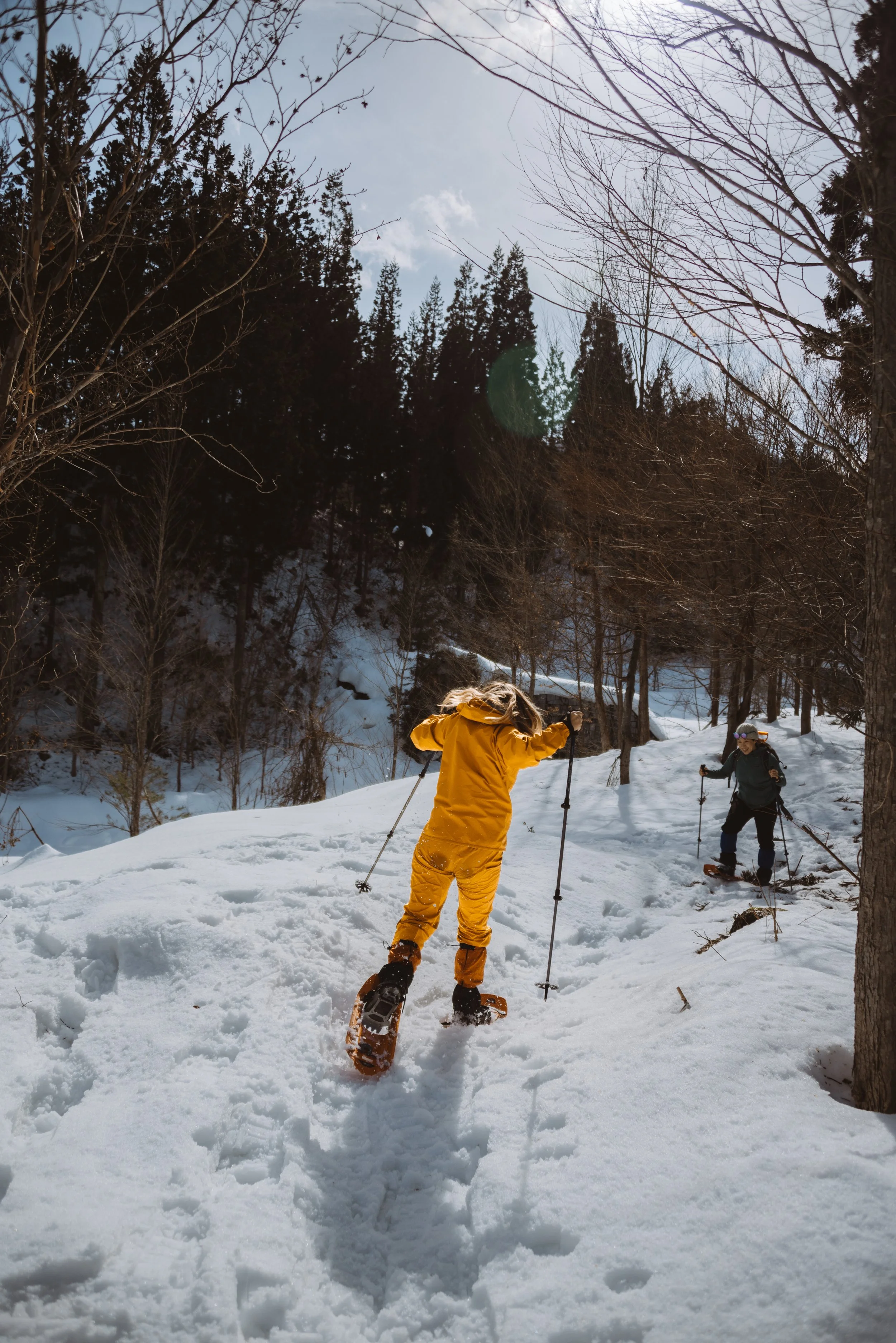 Two children snowshoeing on a snowy trail in a forest with tall trees, one wearing a yellow jacket and pants, the other in dark clothing, under a partly cloudy sky.