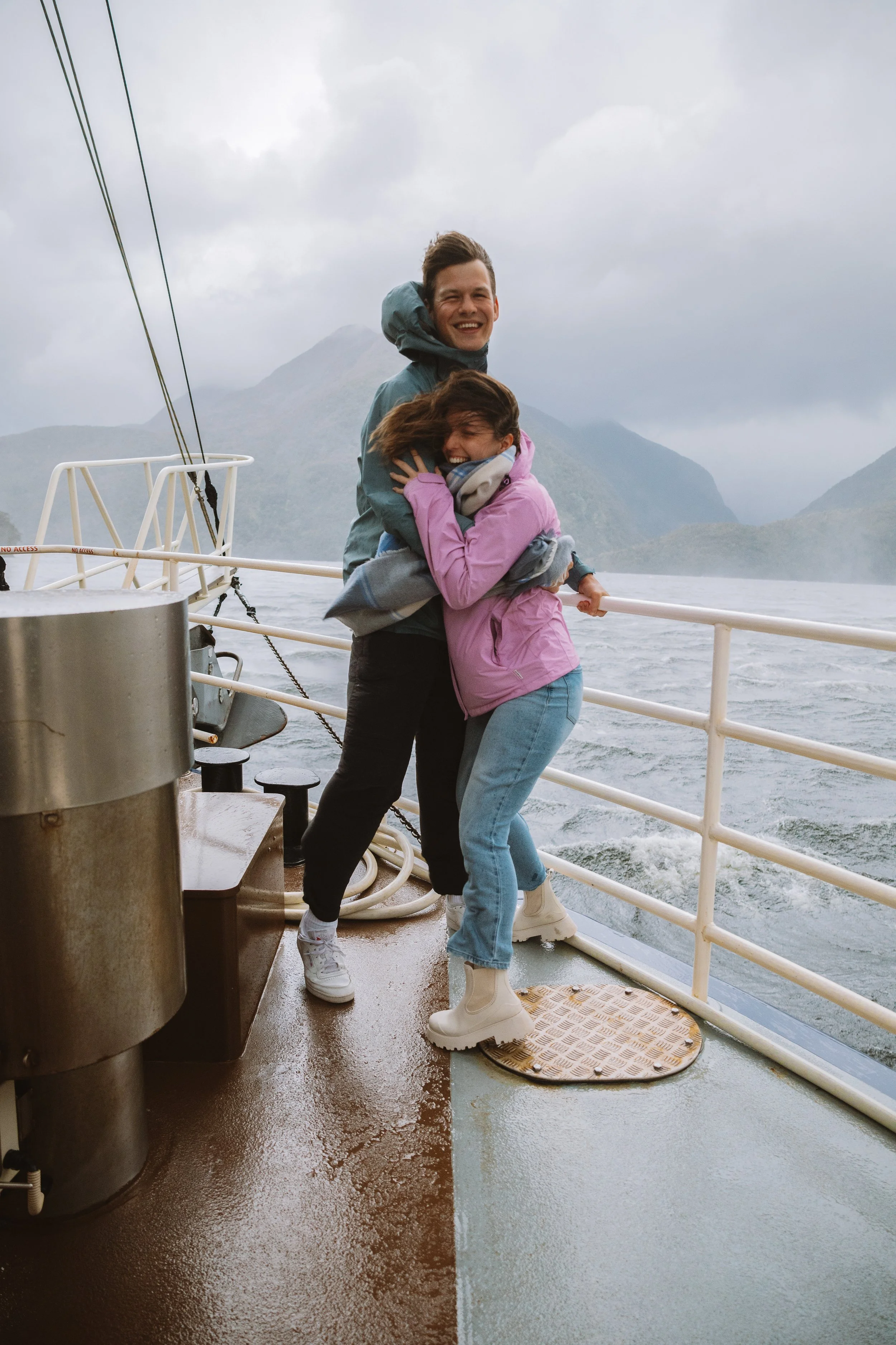 Two people hugging and smiling on a boat deck, with a lake, mountains, and cloudy sky in the background.
