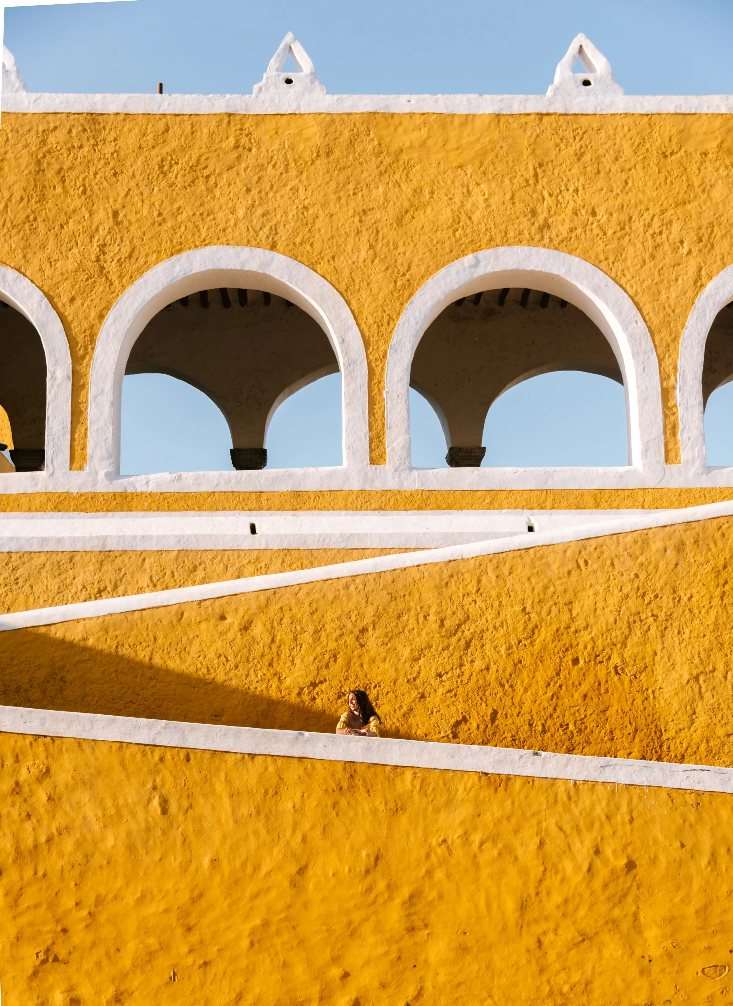 A woman is sitting on a white ledge in front of a yellow textured wall with large white arches and a white upper structure with triangular shapes, against a blue sky.