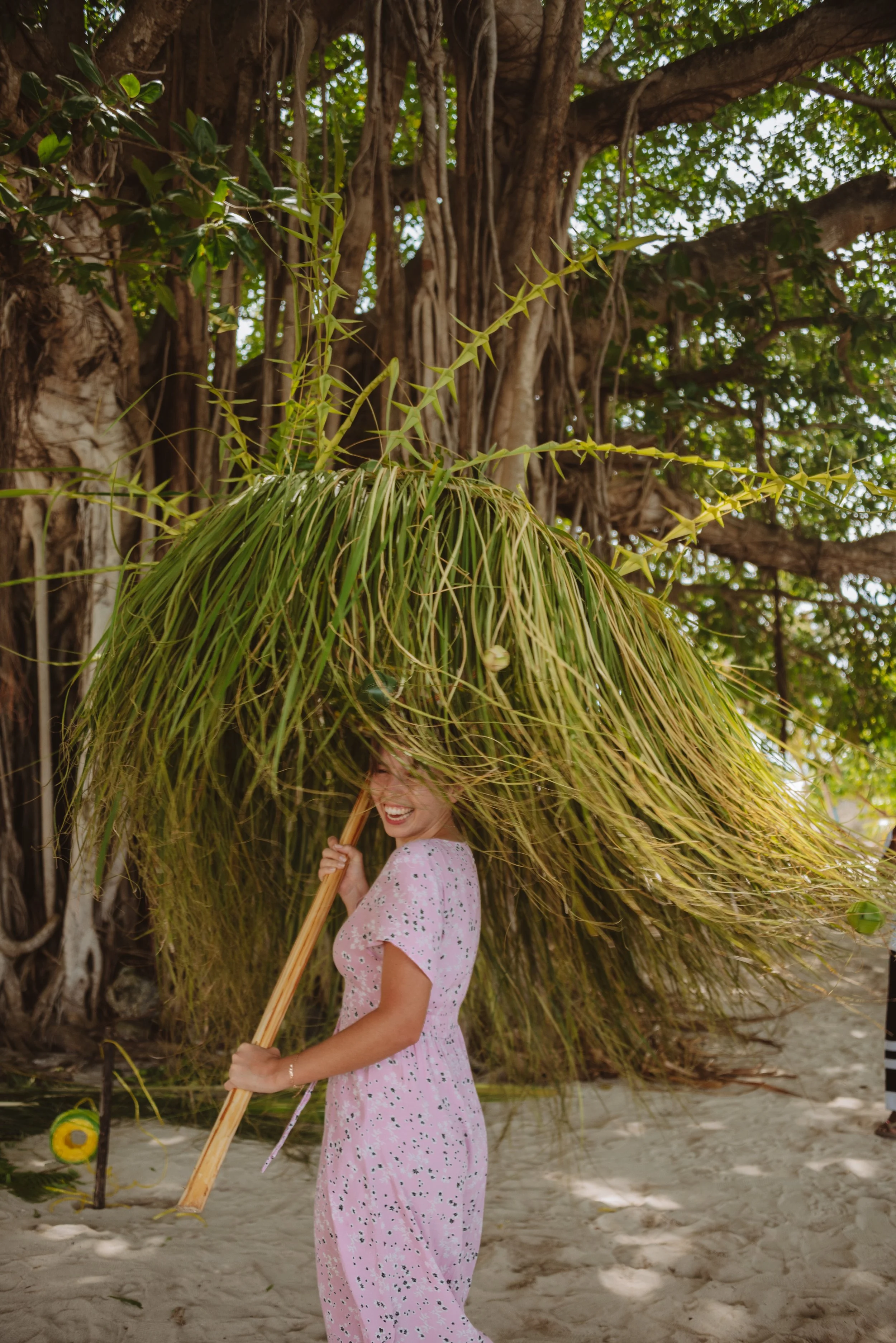 A woman smiling under a large thatched umbrella made of palm or similar leaves, standing on a sandy beach in front of a large tree with thick trunk and green foliage.