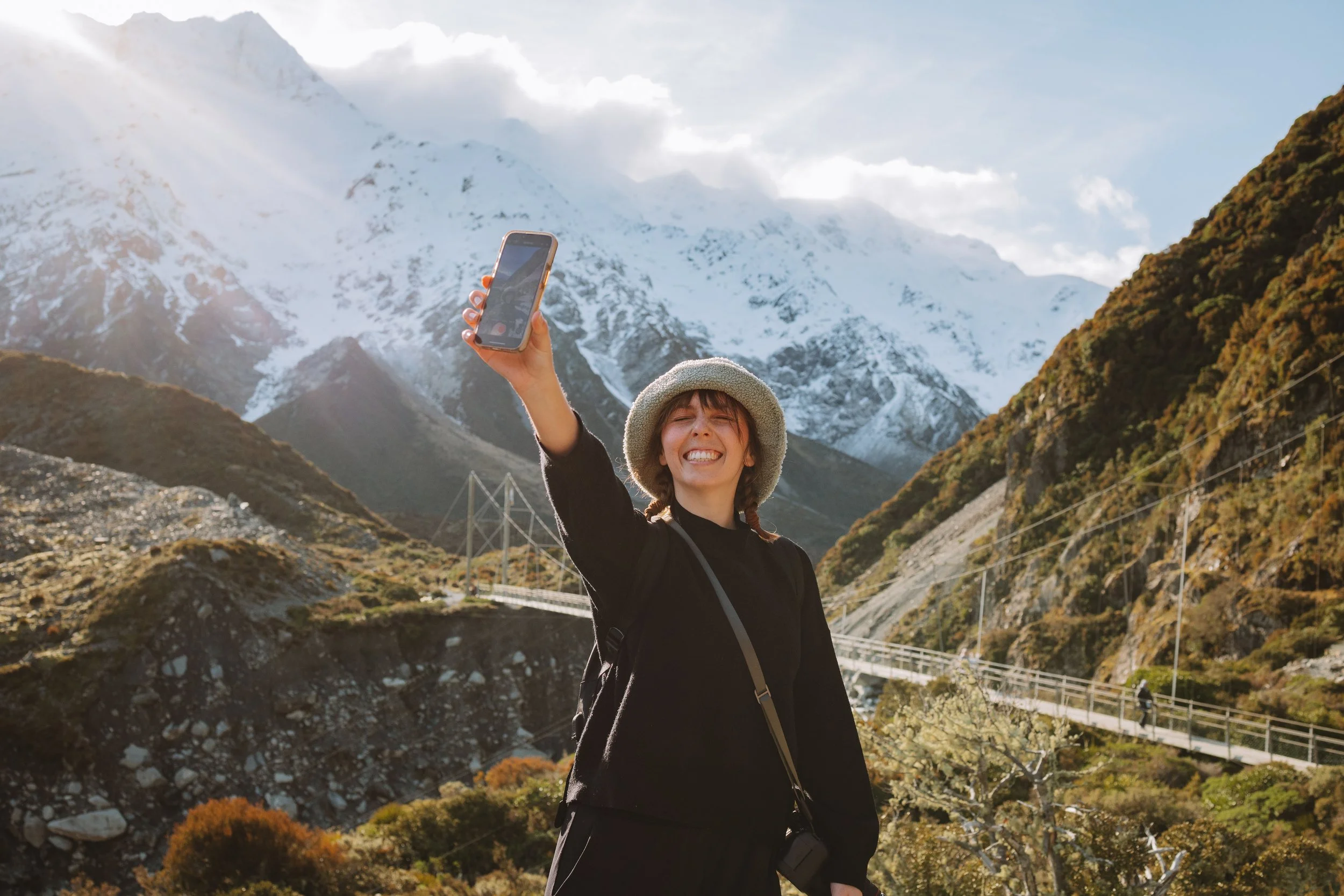 Smiling woman taking a selfie outdoors in mountainous terrain with snow-capped peaks, a bridge, and green hills in the background