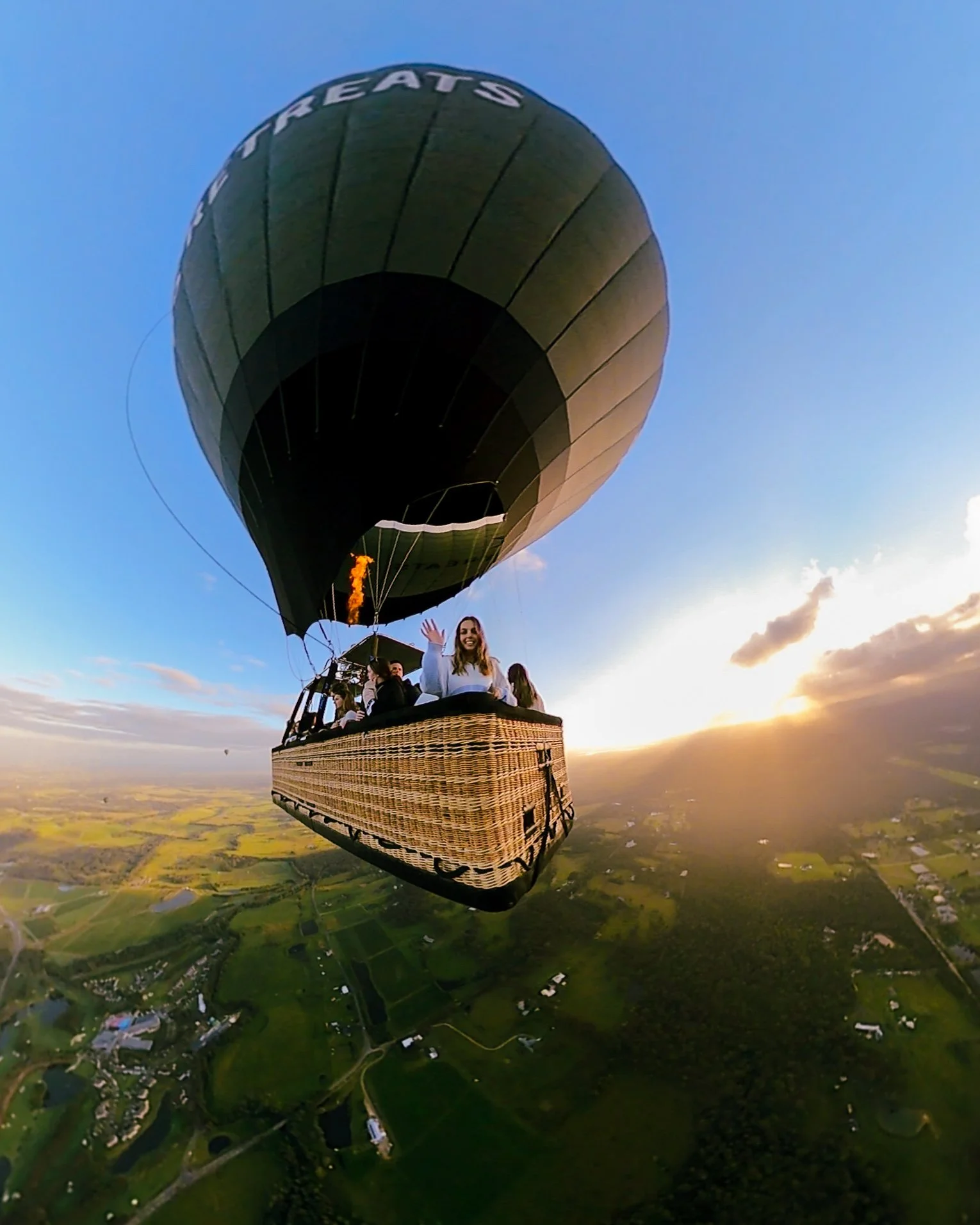 Hot air balloon flying at sunset with passengers, one woman waving, over a green rural landscape.