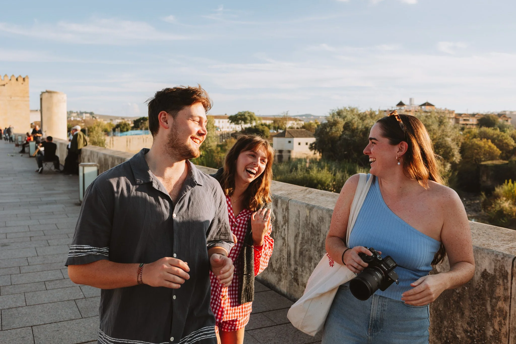 Three people laughing and talking on a bridge with a historic city in the background, during the daytime.