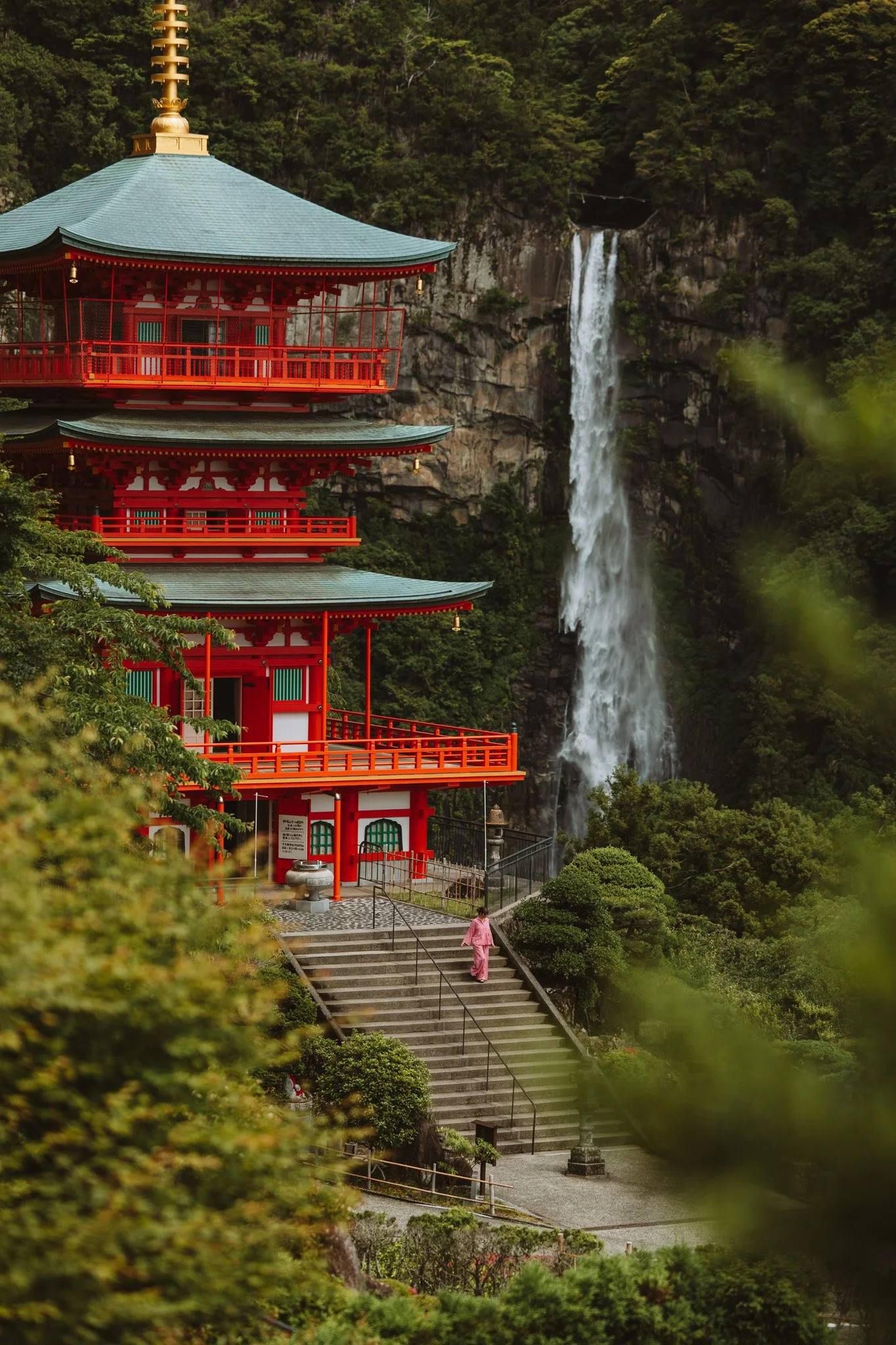 A traditional Japanese pagoda-style building painted in red, situated among lush greenery with a waterfall in the background.