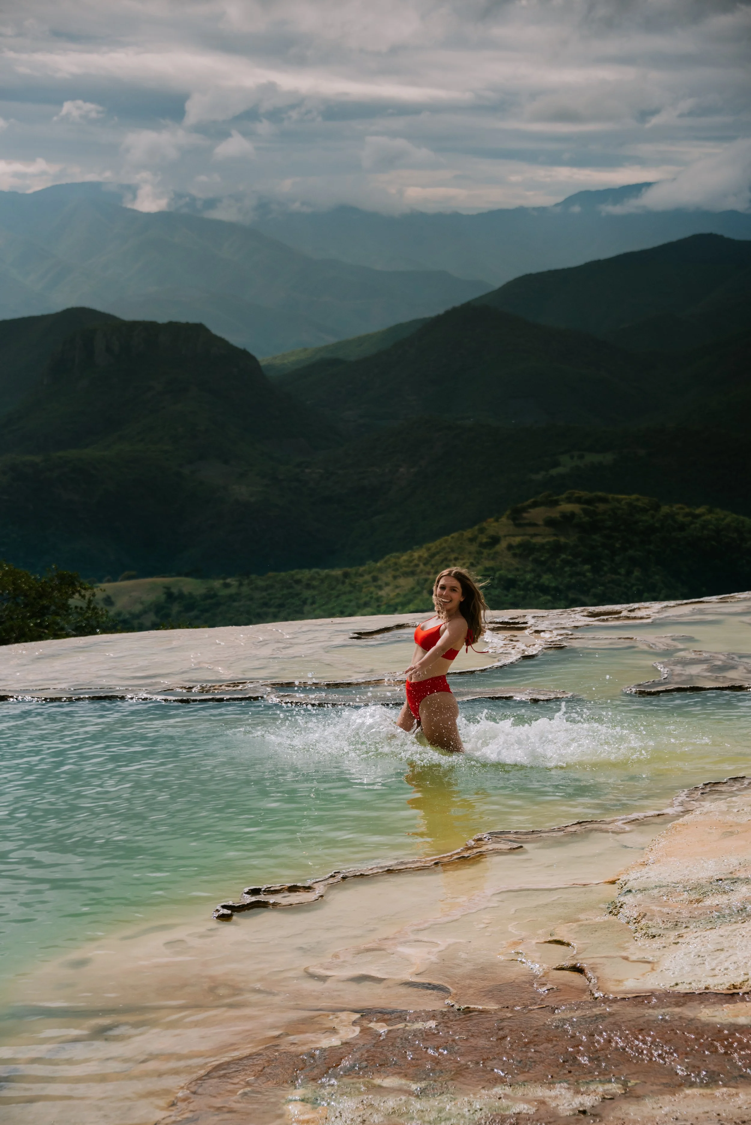 A woman in a red bikini standing in a natural hot spring with mountains in the background.