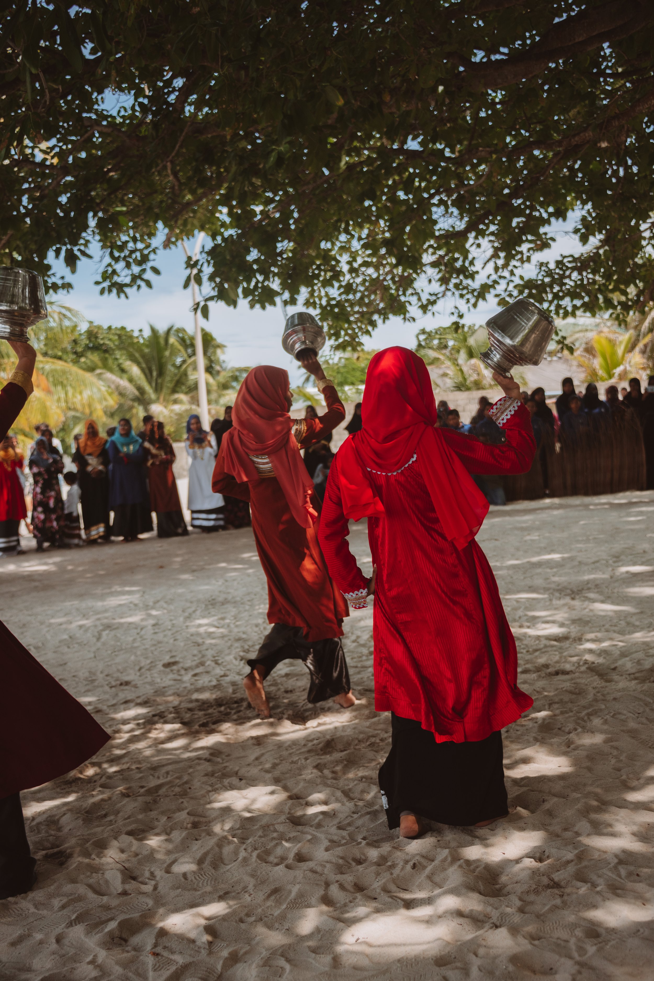 Women holding metal containers on their heads in a sandy outdoor area, with people standing in the background under a tree.