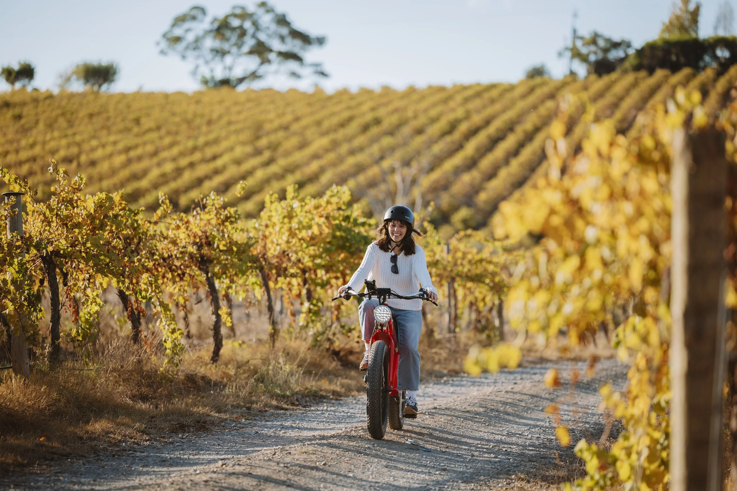 A woman riding a mountain bike on a dirt path through a vineyard during fall, wearing a helmet and sunglasses, smiling amidst the golden leaves.
