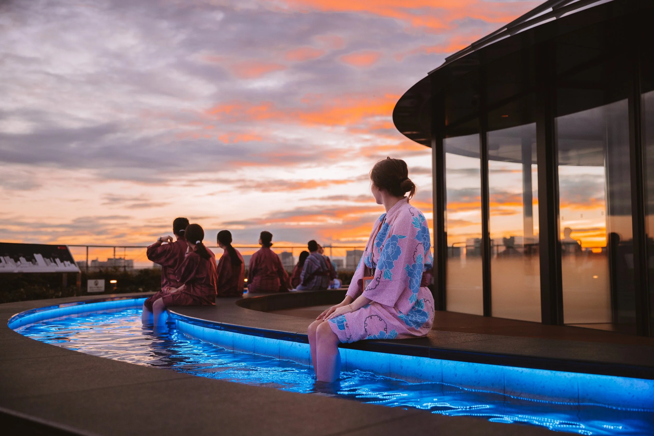 People in traditional kimonos sitting and standing near a blue-lit hot spring pool at sunset, with a modern building and city skyline in the background.