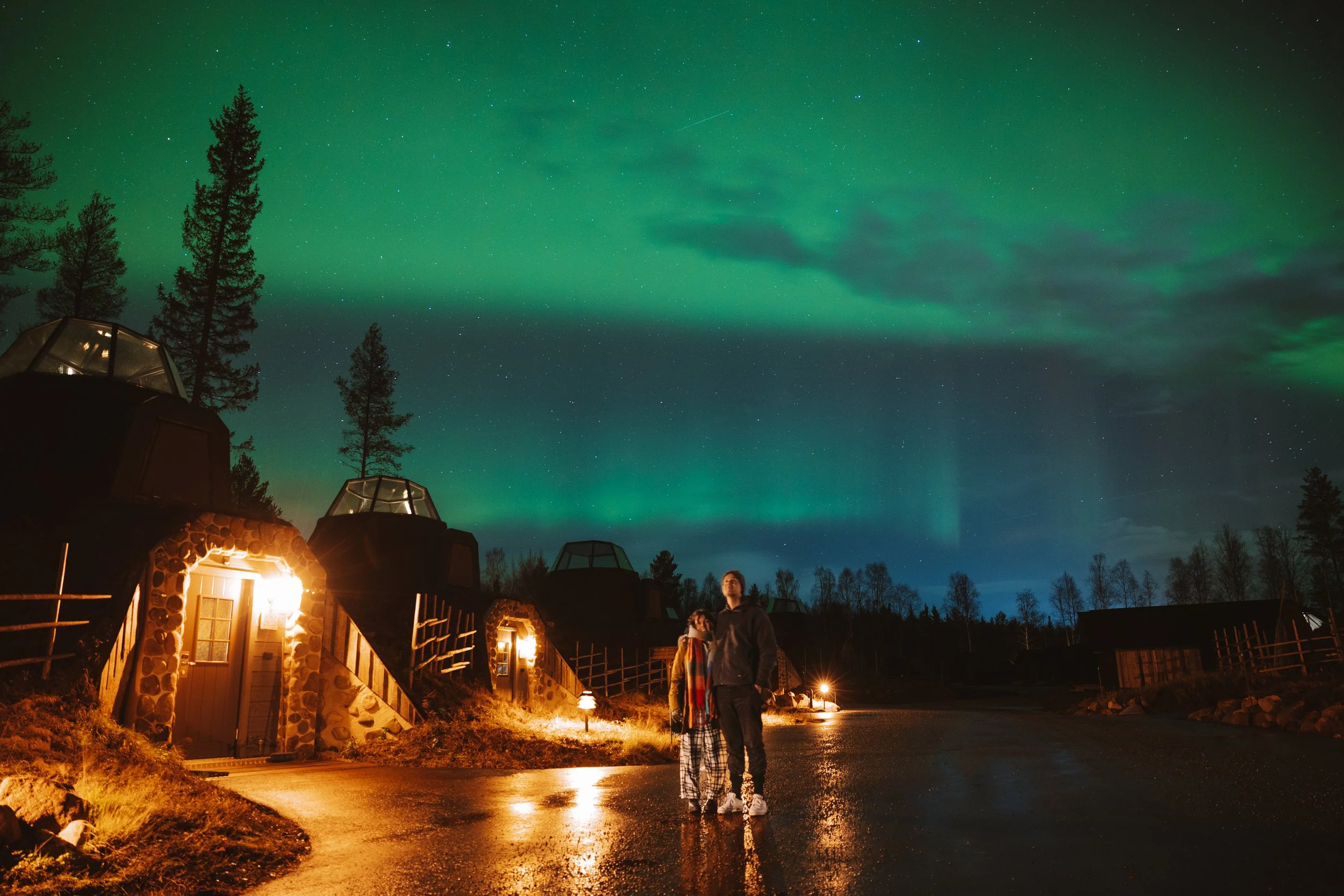 Two people standing on a wet street at night with the Northern Lights visible in the sky above. Small buildings with lights are on either side, and tall trees are in the background.