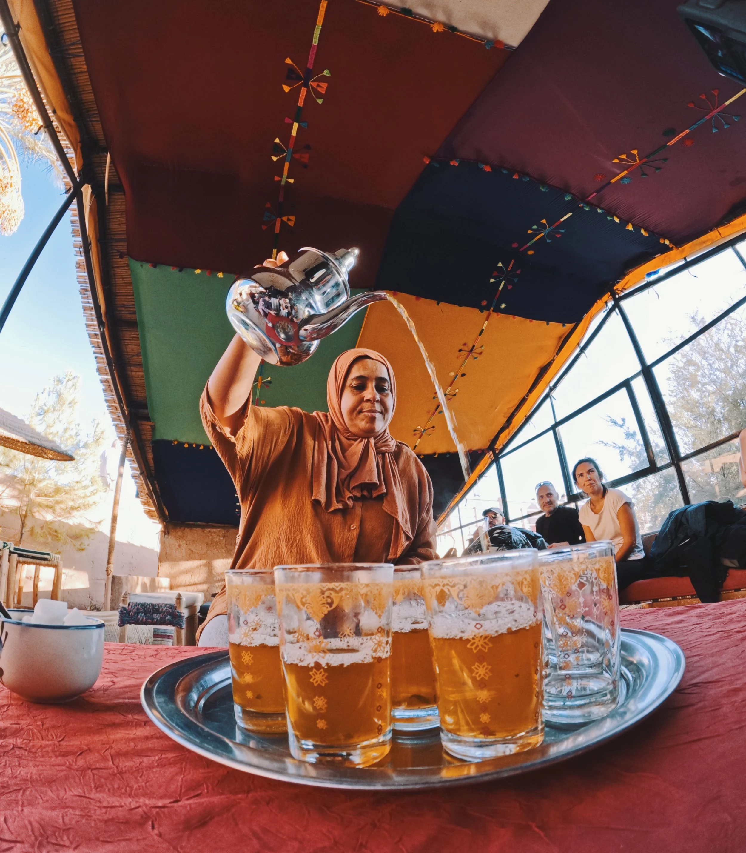 A woman wearing a brown headscarf and matching top pouring water into glasses of beer on a tray, with a group of people sitting in the background under a colorful canopy.
