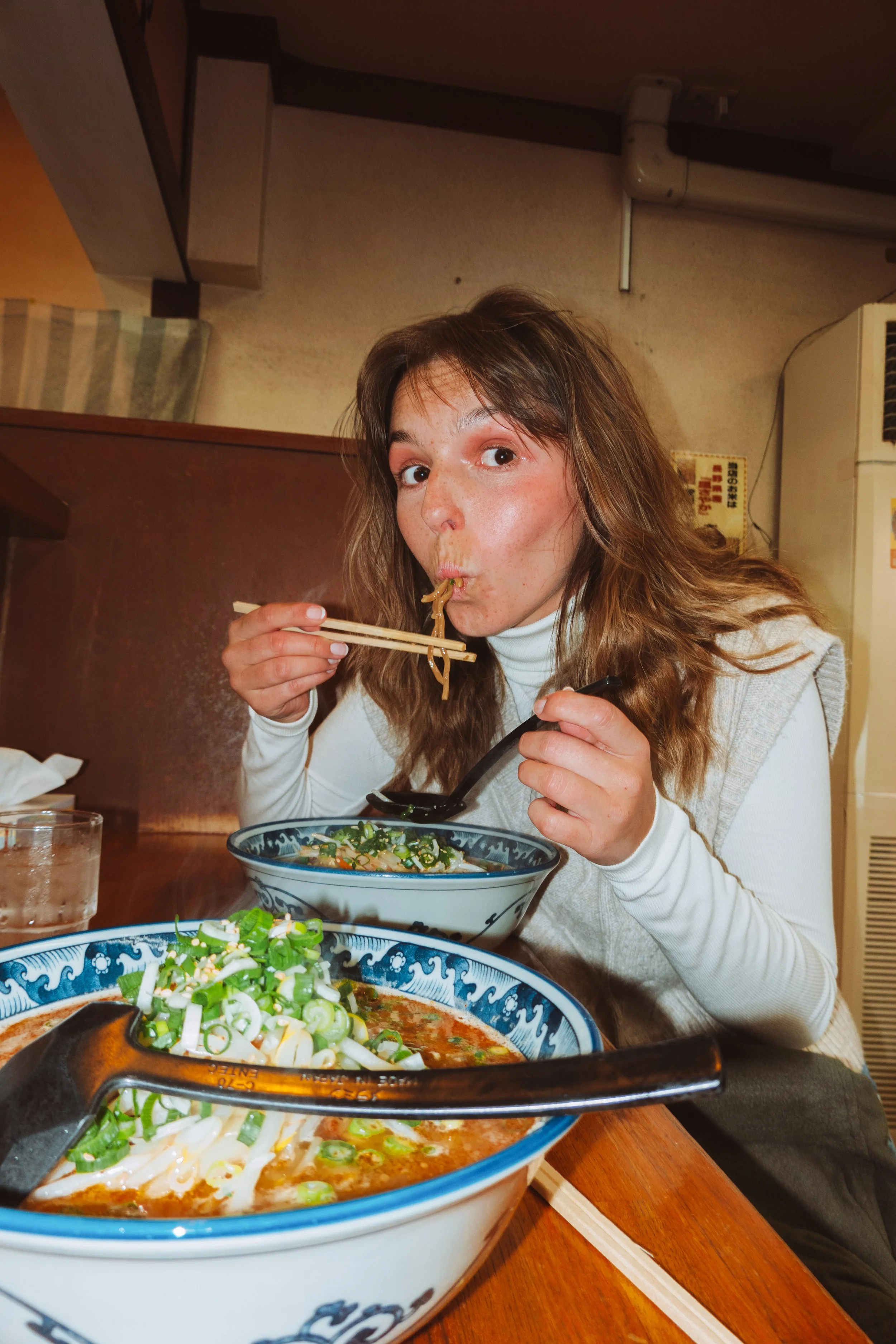A woman with long wavy brown hair eating ramen noodles with chopsticks in a restaurant. There are two bowls of ramen with green onions on the table, one in front of her and one near the front of the image, along with a glass of water.