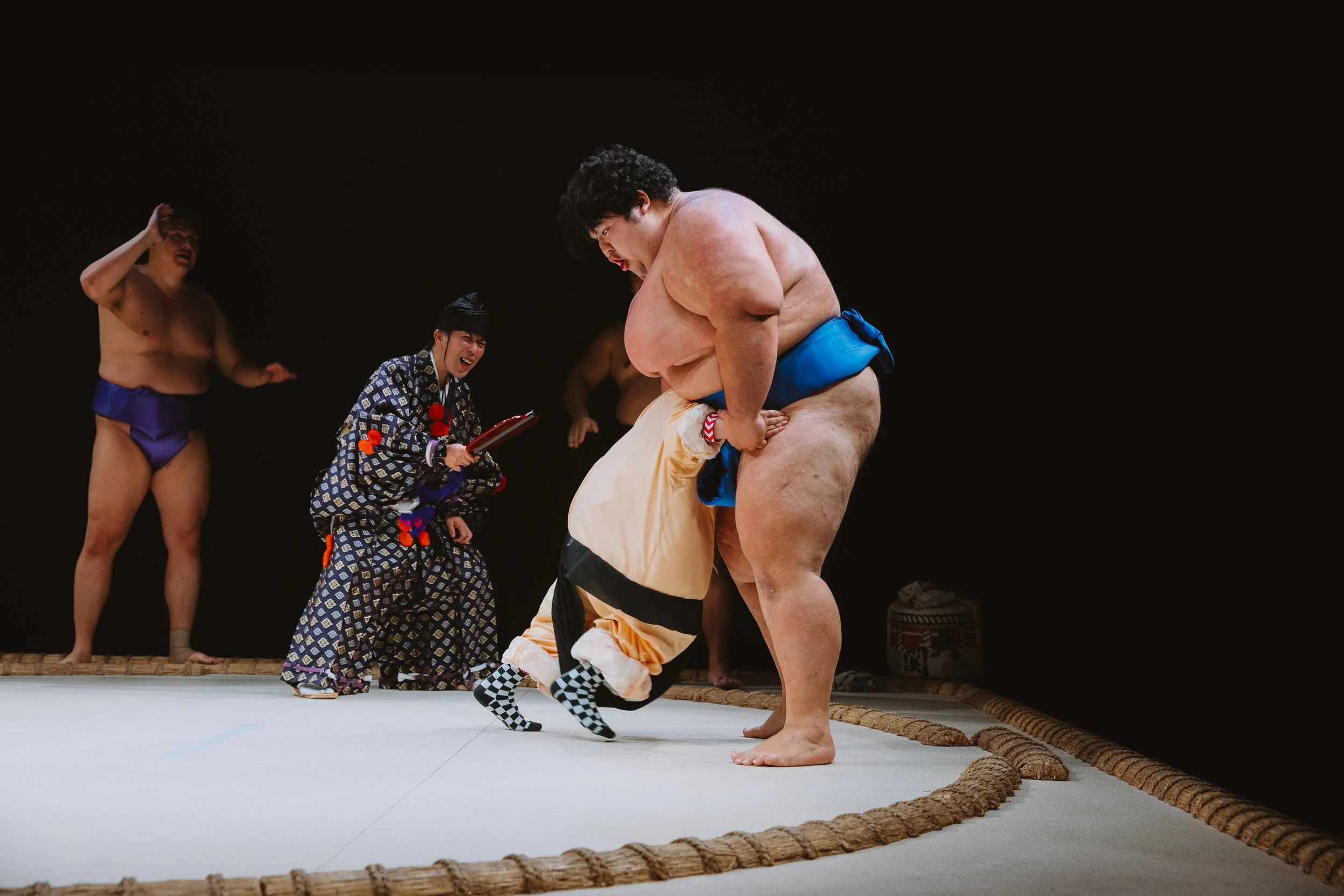 Sumo wrestlers fighting in a sumo ring, with referees and attendants watching on.