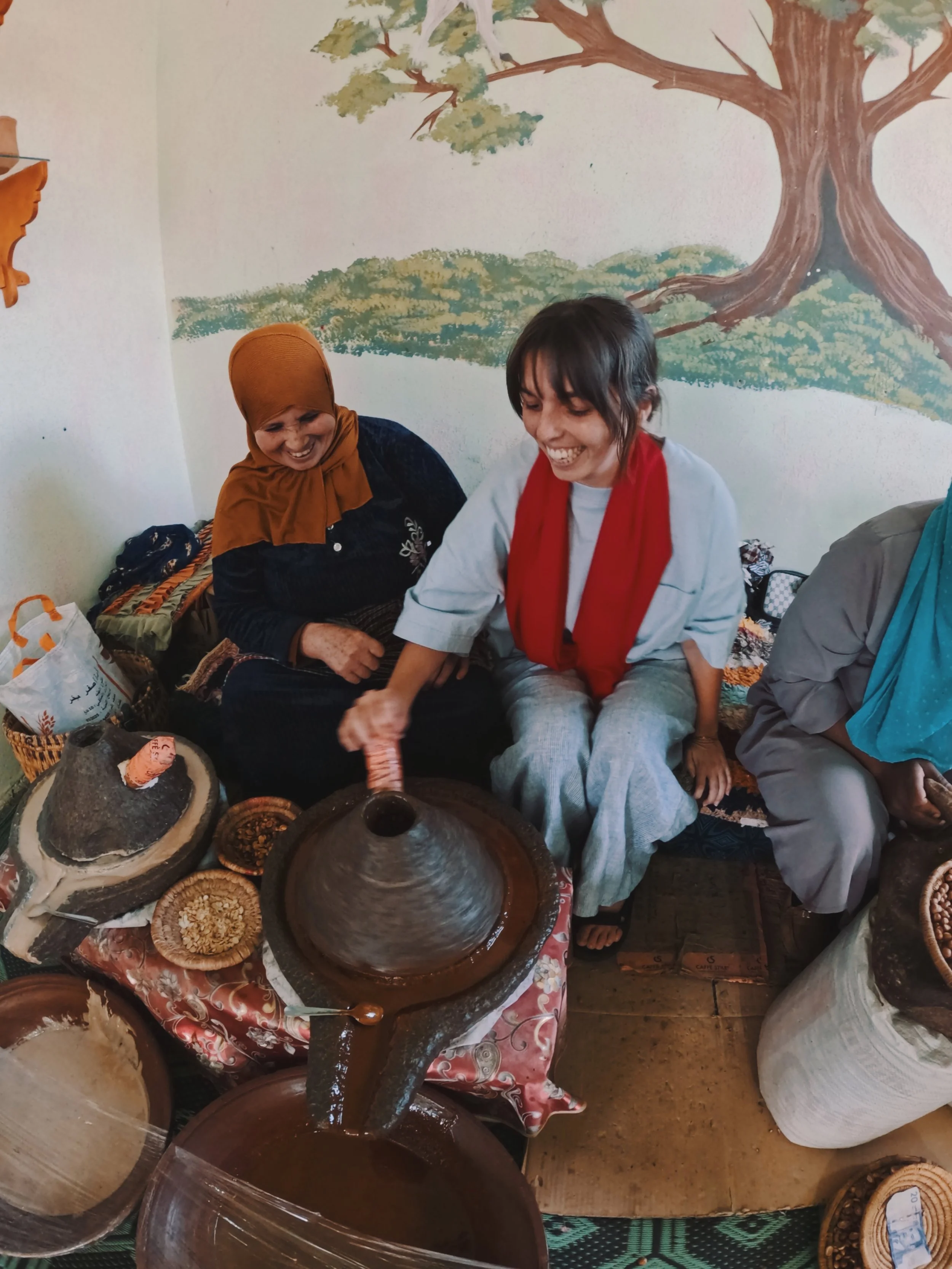 Two women are sitting at a table, grinding spices or herbs using traditional tools, and smiling. They are in a room with a painted tree mural on the wall behind them, and various bowls and containers of spices or ingredients are on the table.