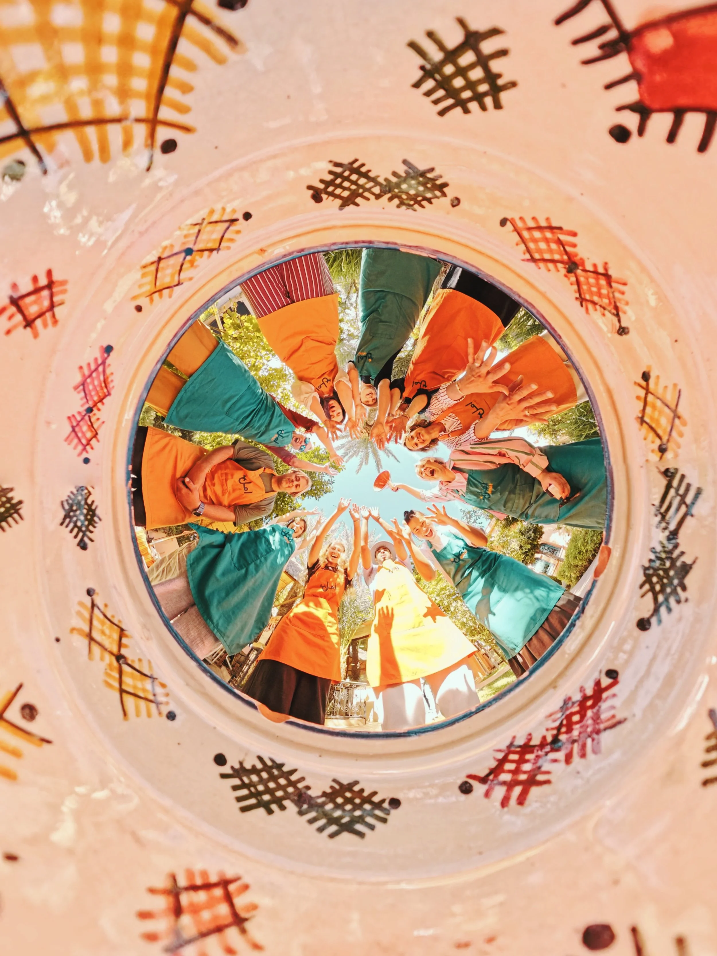 People in colorful outfits are gathered outdoors, raising their hands and smiling, viewed from inside a decorated circular object.