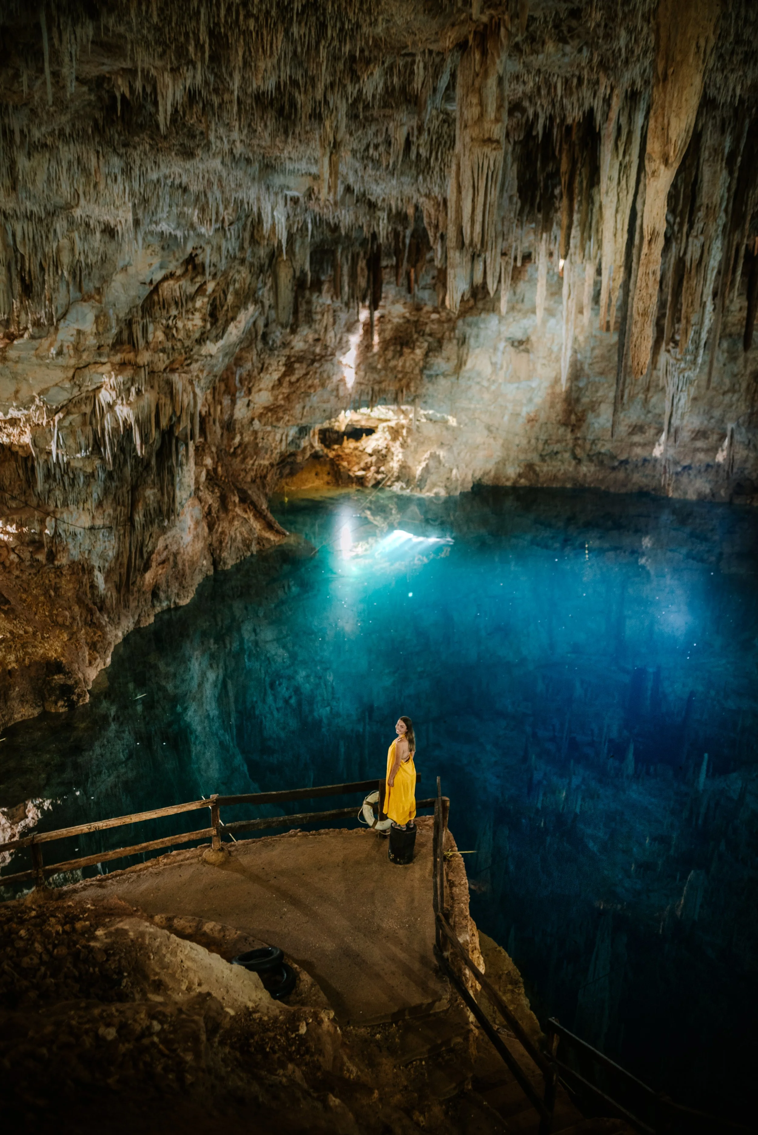 A woman in a yellow dress standing by a railing inside a large underground cave with a deep blue lake and stalactites hanging from the ceiling.
