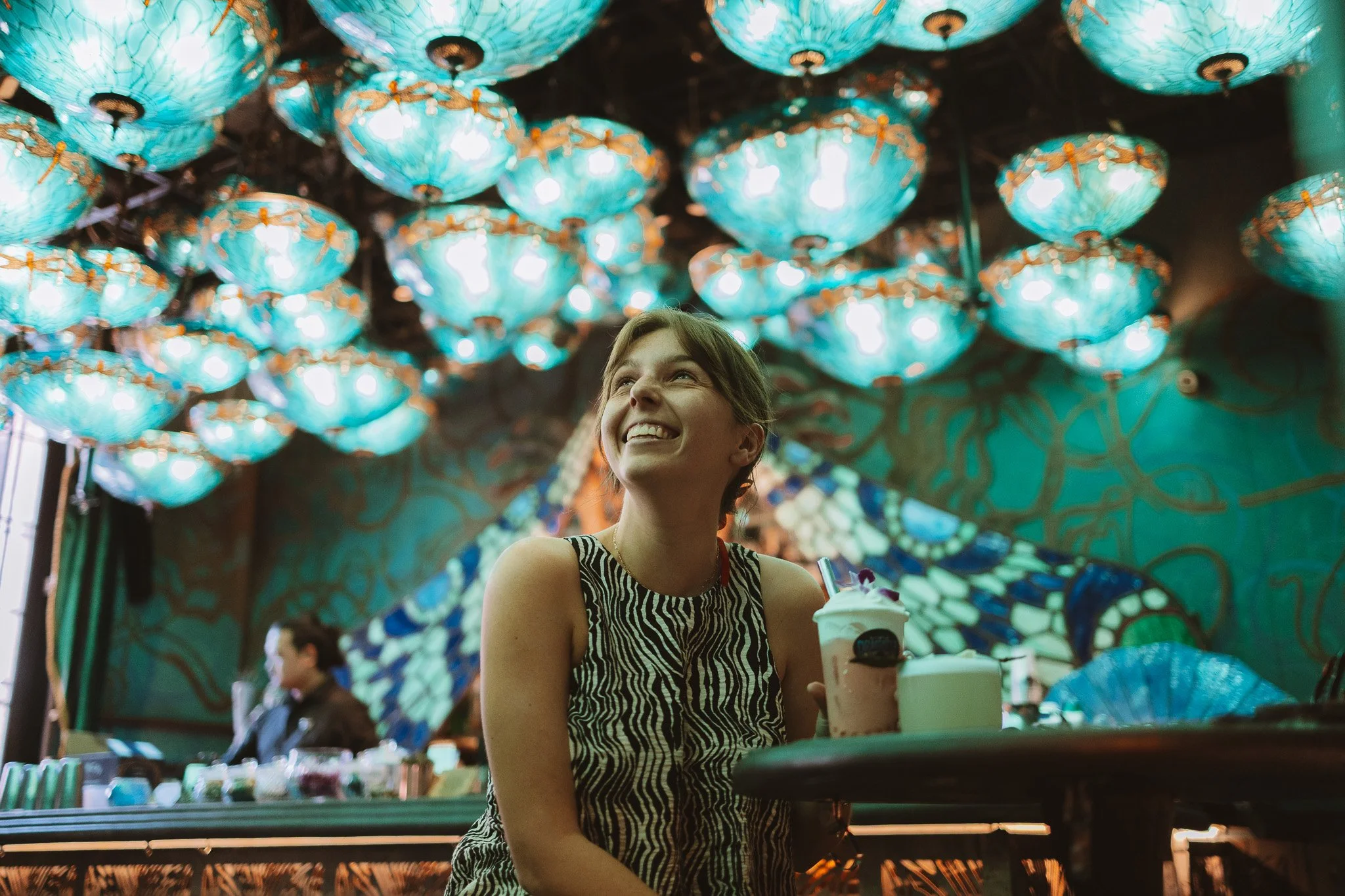 A young woman with short hair, smiling and looking up, sitting at a table in a dimly lit space decorated with vibrant, glowing blue lanterns and a mural of a butterfly on the wall.