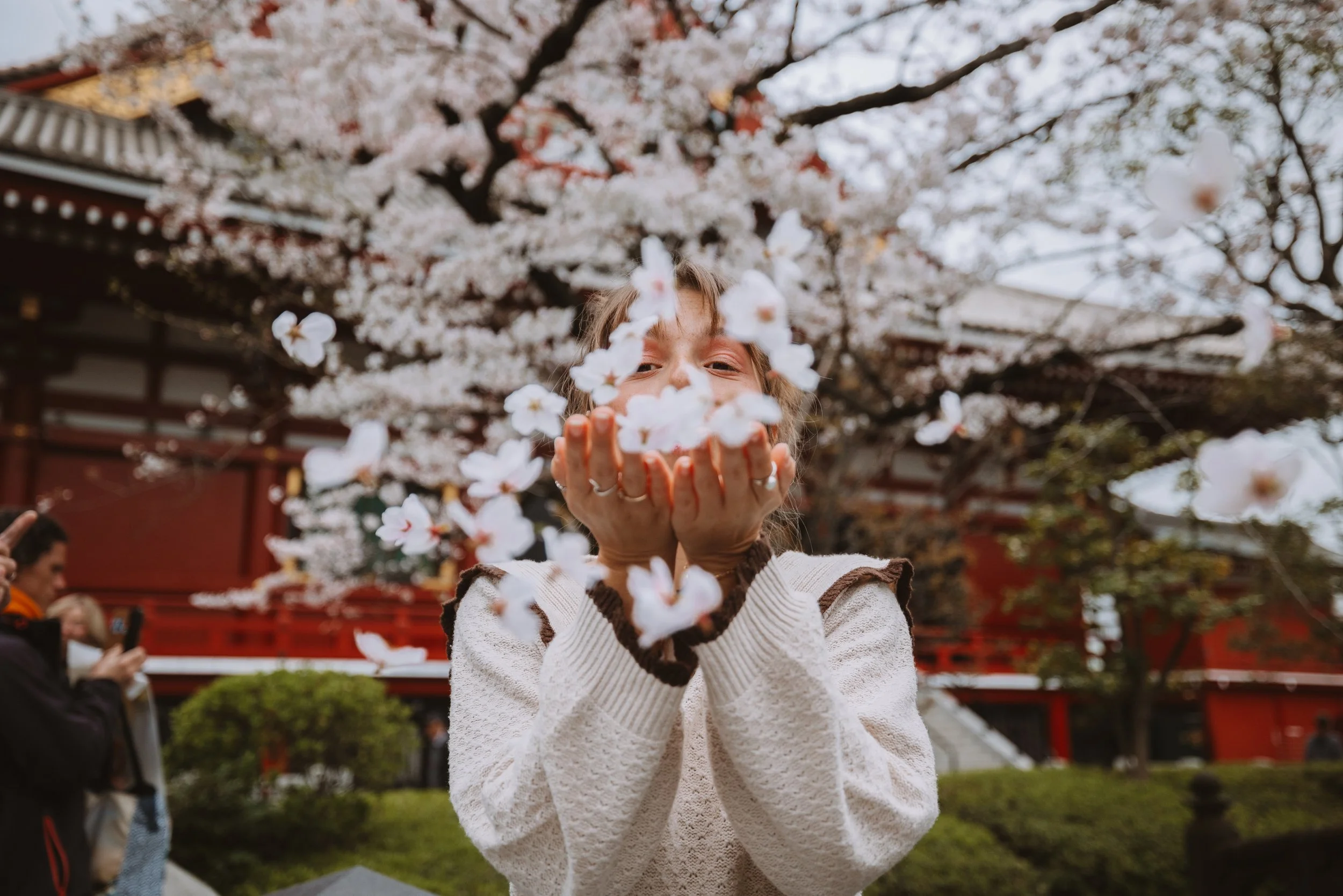 Woman blowing cherry blossom petals in front of traditional Japanese temple.