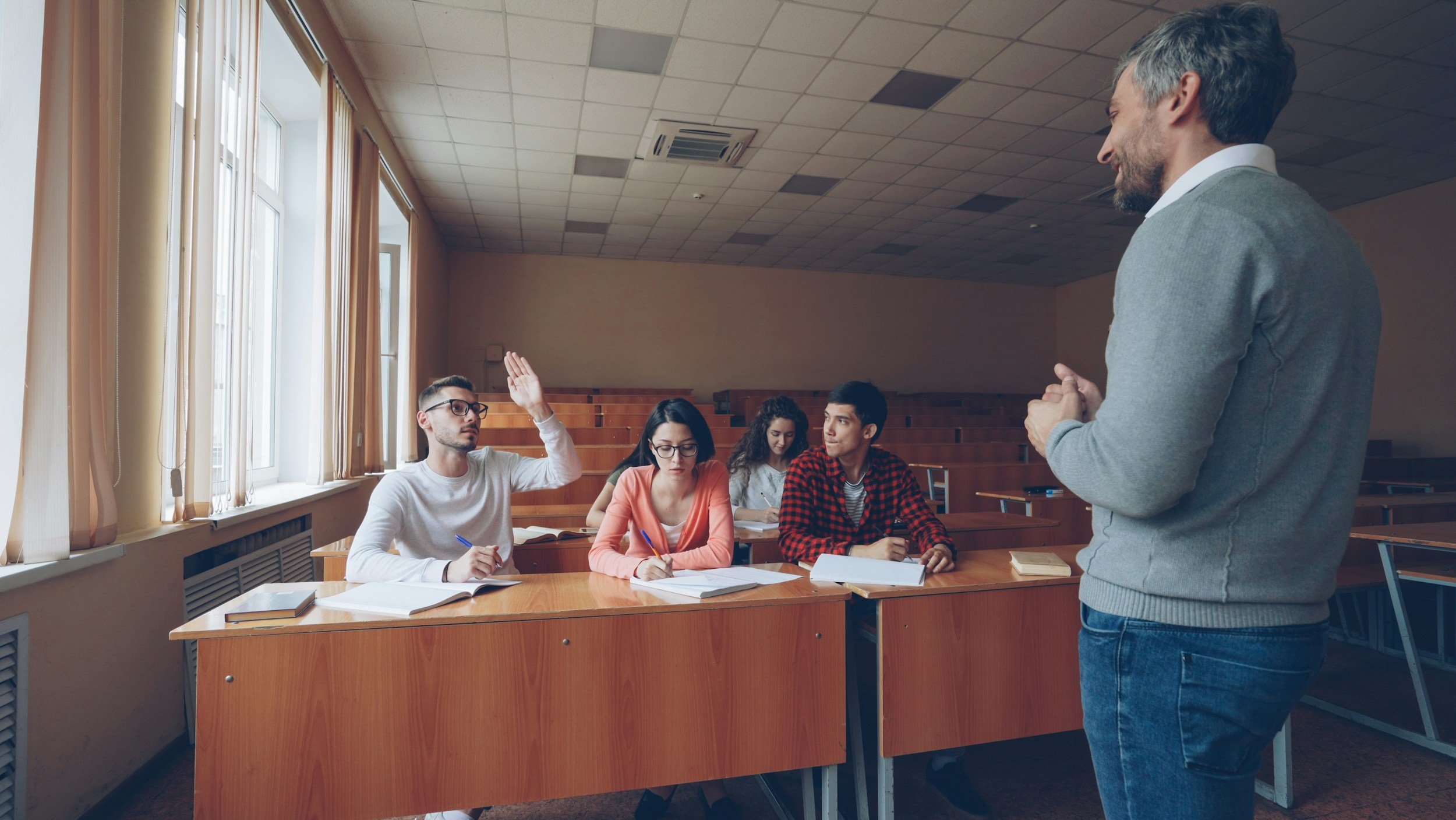 A classroom scene with five students seated at wooden desks, with a teacher standing at the front. One student raises his hand, while others take notes or look at the teacher. The classroom has large windows with curtains and a tiled ceiling.