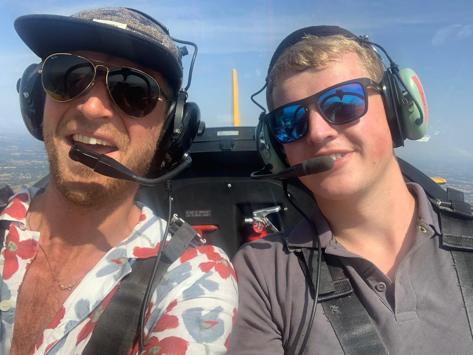 Paul Reynolds and pioneer Sam Mayall taking a selfie in a small aircraft with a clear sky and landscape visible in the background, both wearing sunglasses and headsets.