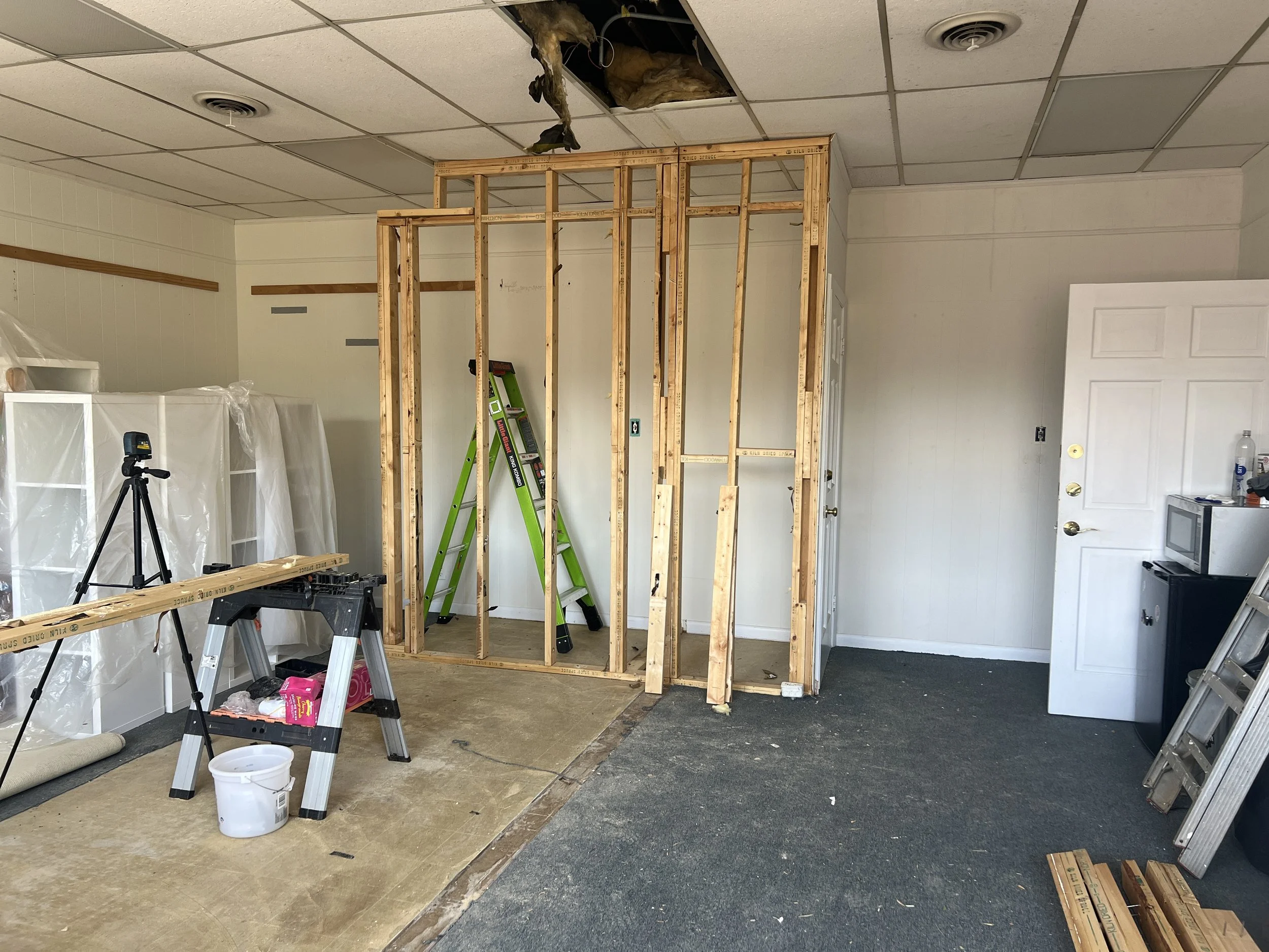 Room under renovation with partially built wooden wall frame, ladder, tools, and construction materials. Ceiling tiles removed, exposing insulation and ceiling ducts.