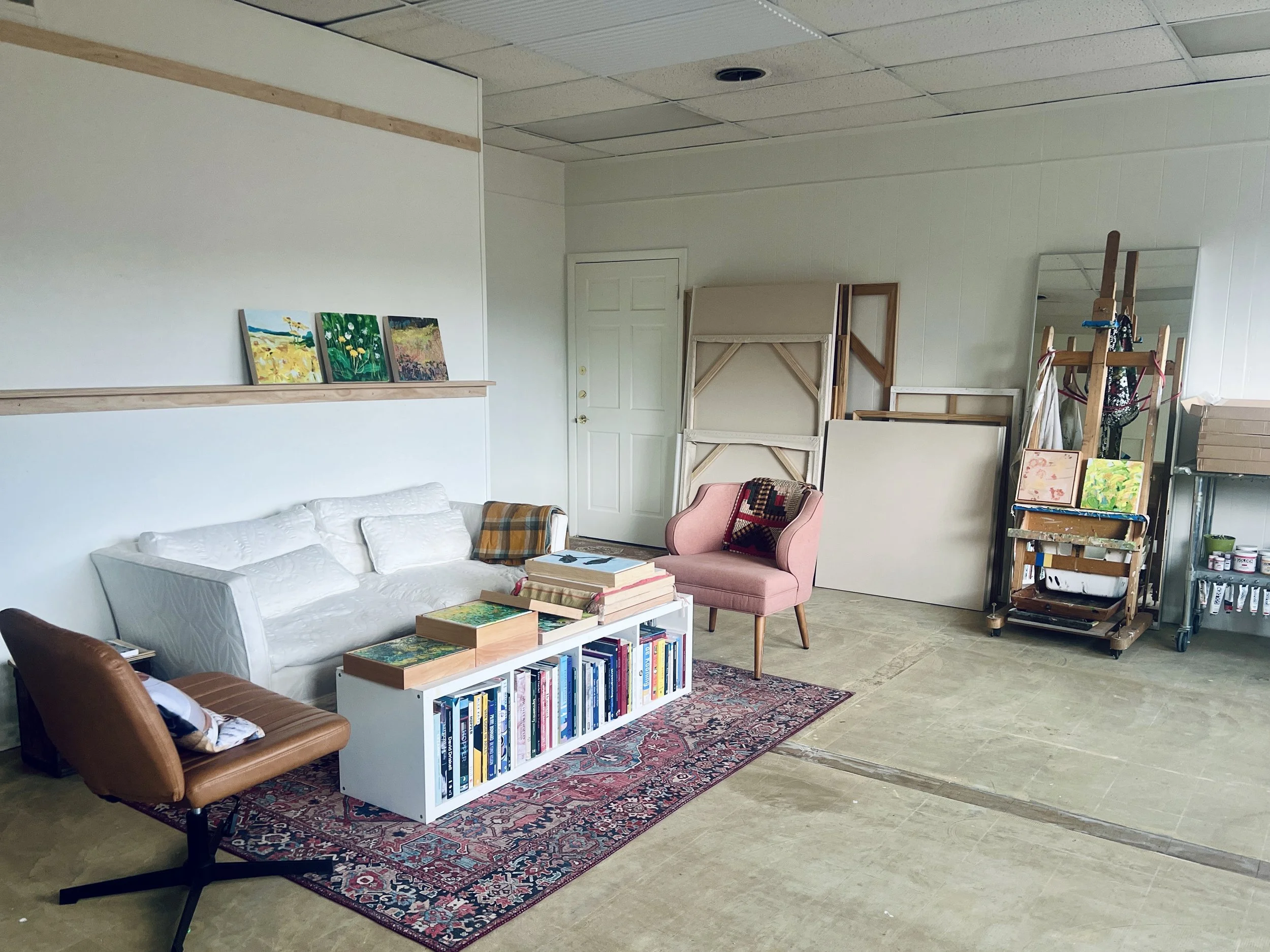 The image displays an art studio or gallery space with a white couch, pink armchair, and brown leather chair arranged around a white bookshelf filled with books. Artwork is propped against the wall, and various art supplies and furniture are scattere