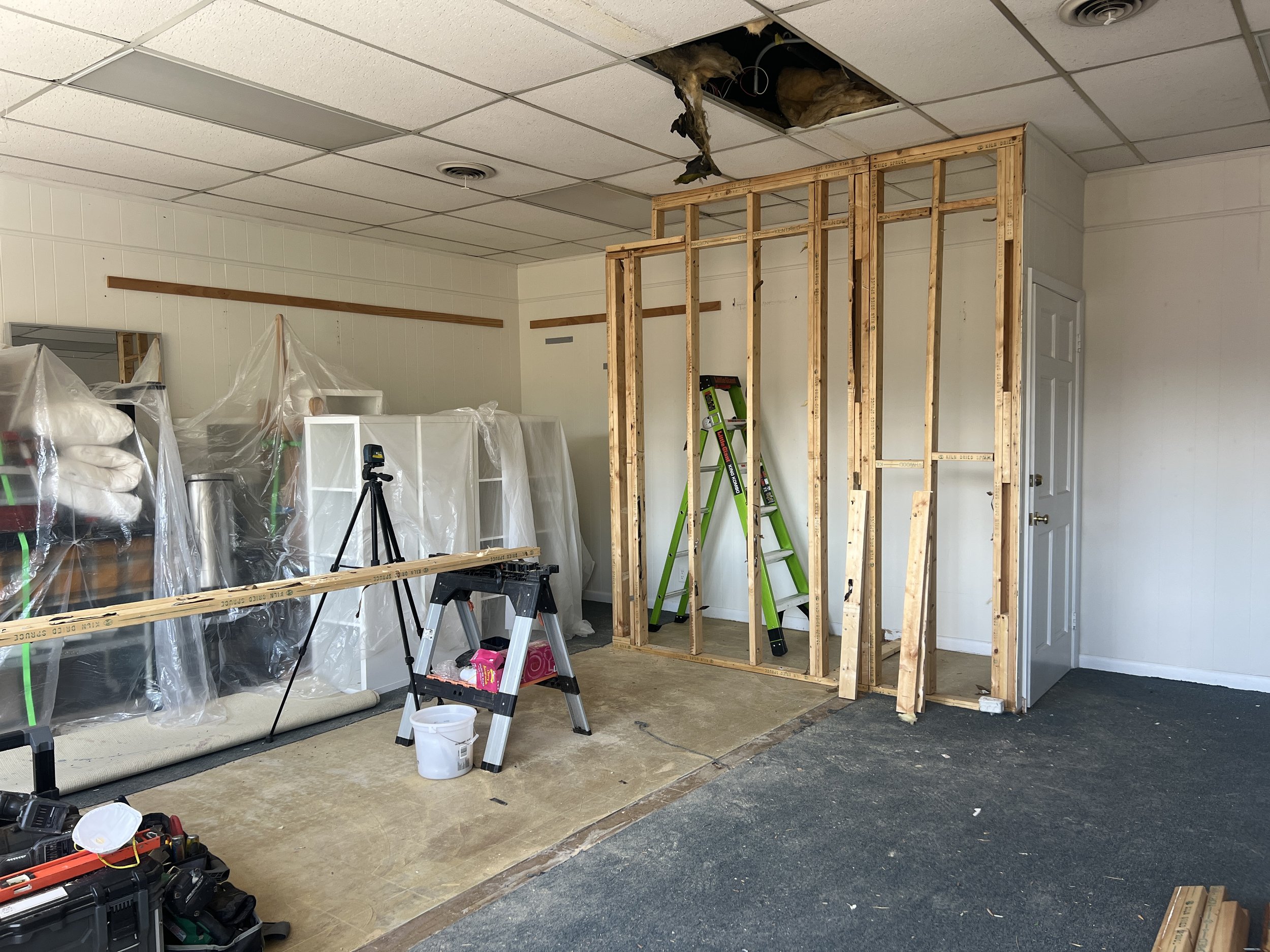 Interior of a room under remodeling with exposed wooden wall studs, construction tools, and plastic-covered furniture. The ceiling has a damaged ceiling tile with insulation hanging down.