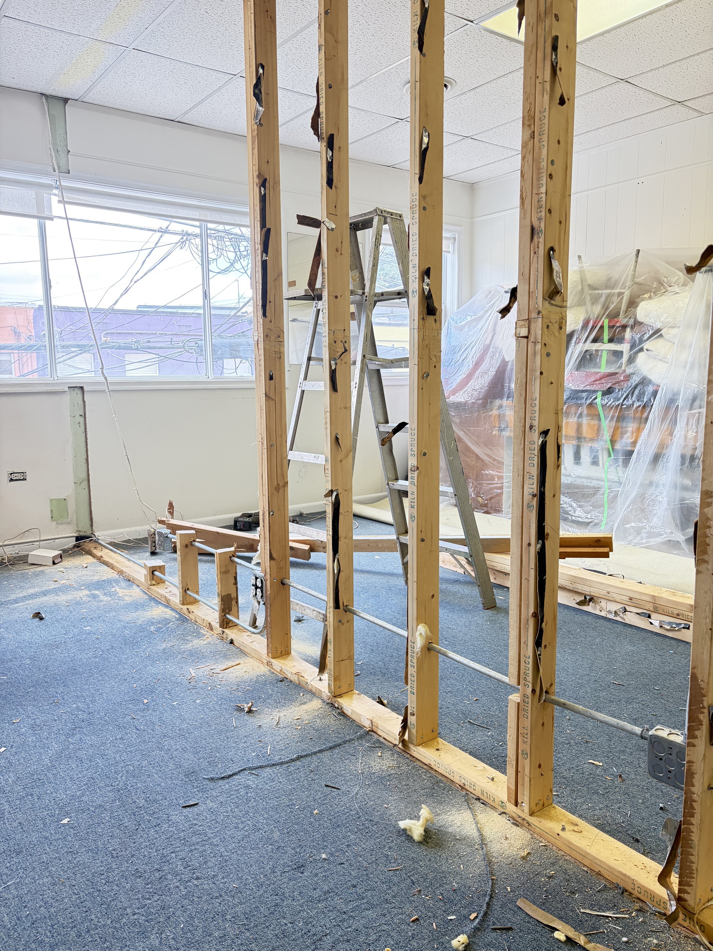 Partially demolished interior wall with exposed wooden studs and electrical wiring, with a ladder and plastic-covered furniture in the background, and debris on the blue carpeted floor.