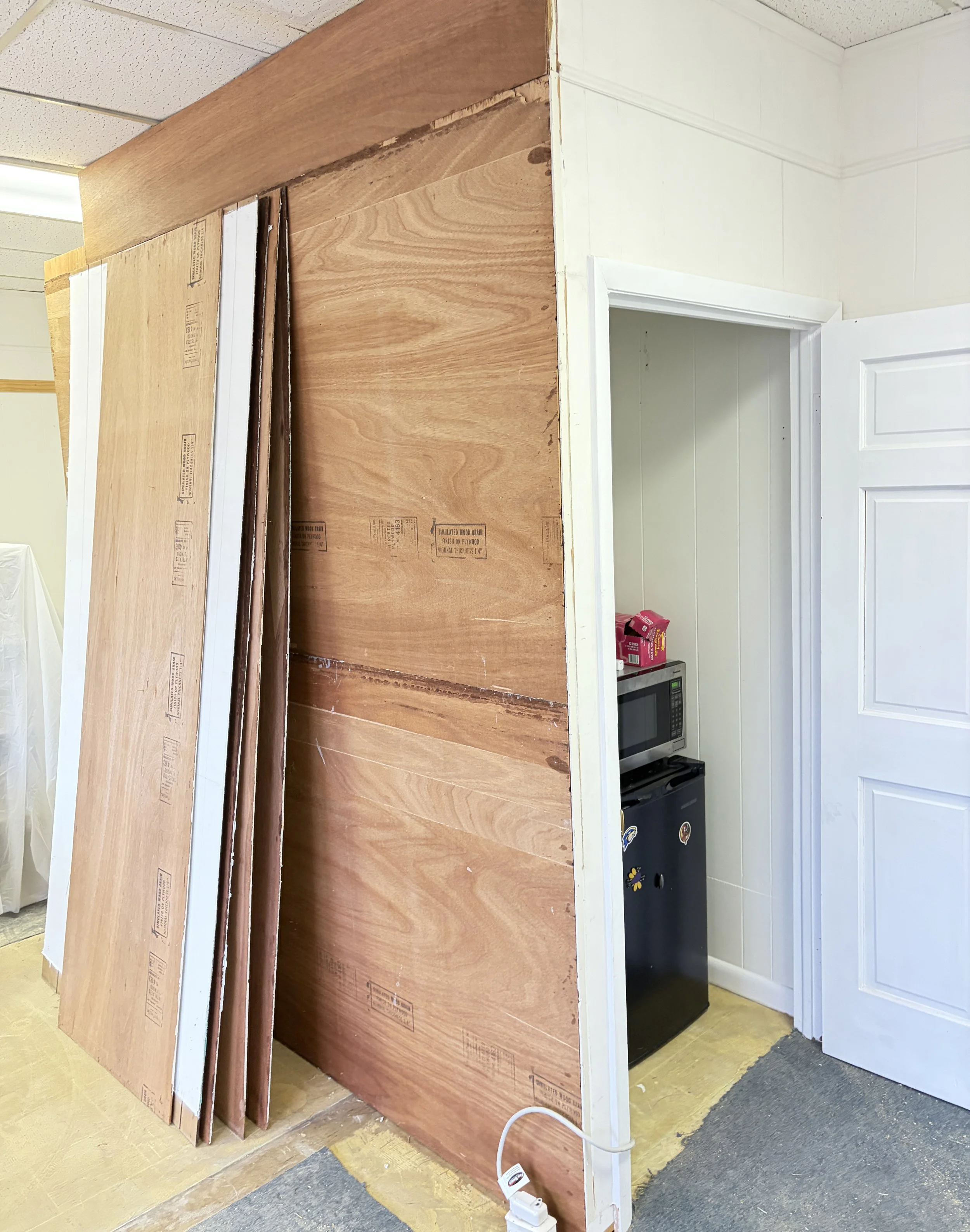 The image shows a partially constructed wall with several large plywood sheets leaning against it, next to an open doorway. Inside the doorway, there is a small black refrigerator and a microwave on top of it. The room has a white door and light-colo