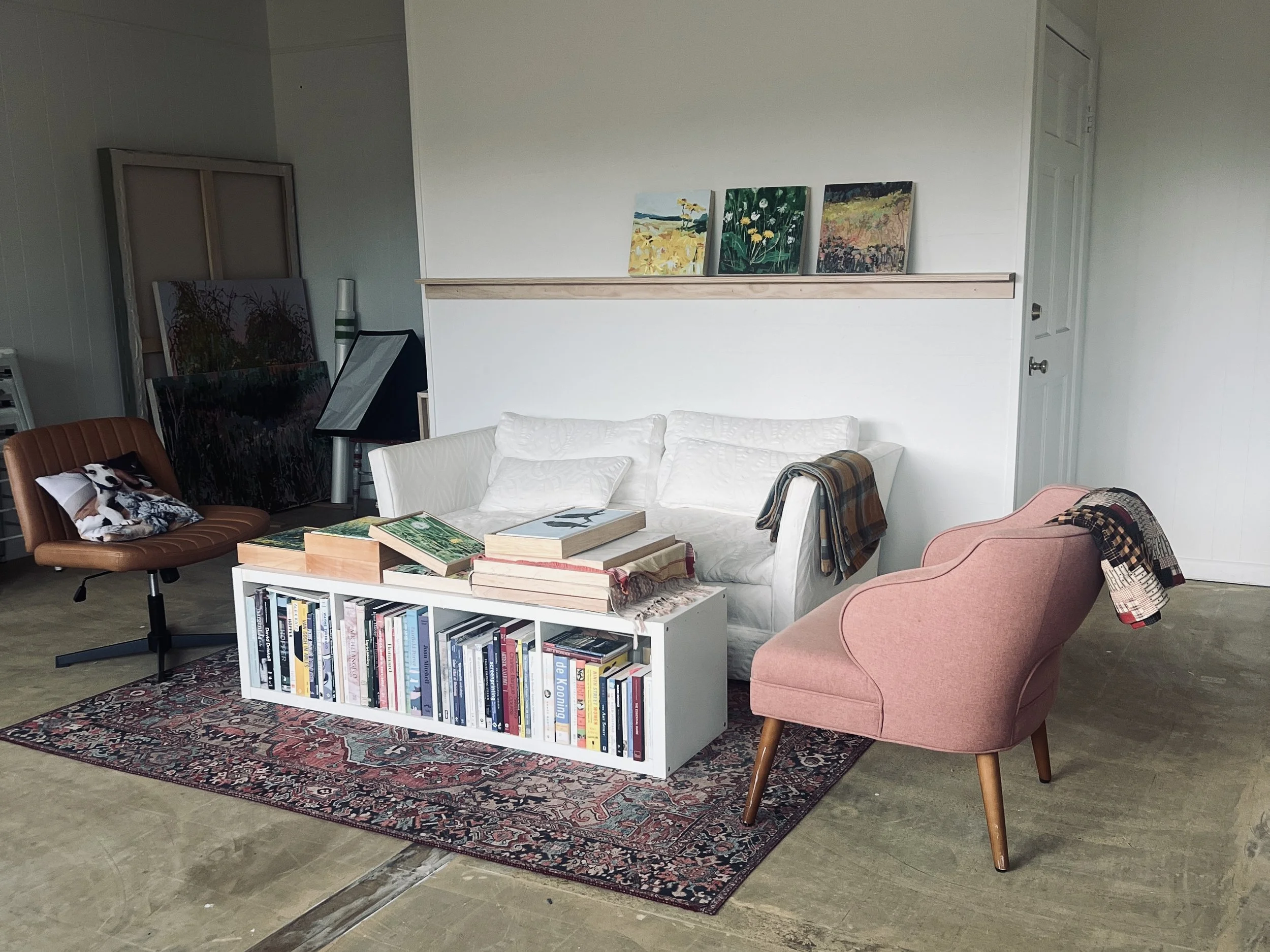 Art studio with white couch, pink armchair, wooden chair, bookshelf, paintings on the wall, and area rug.