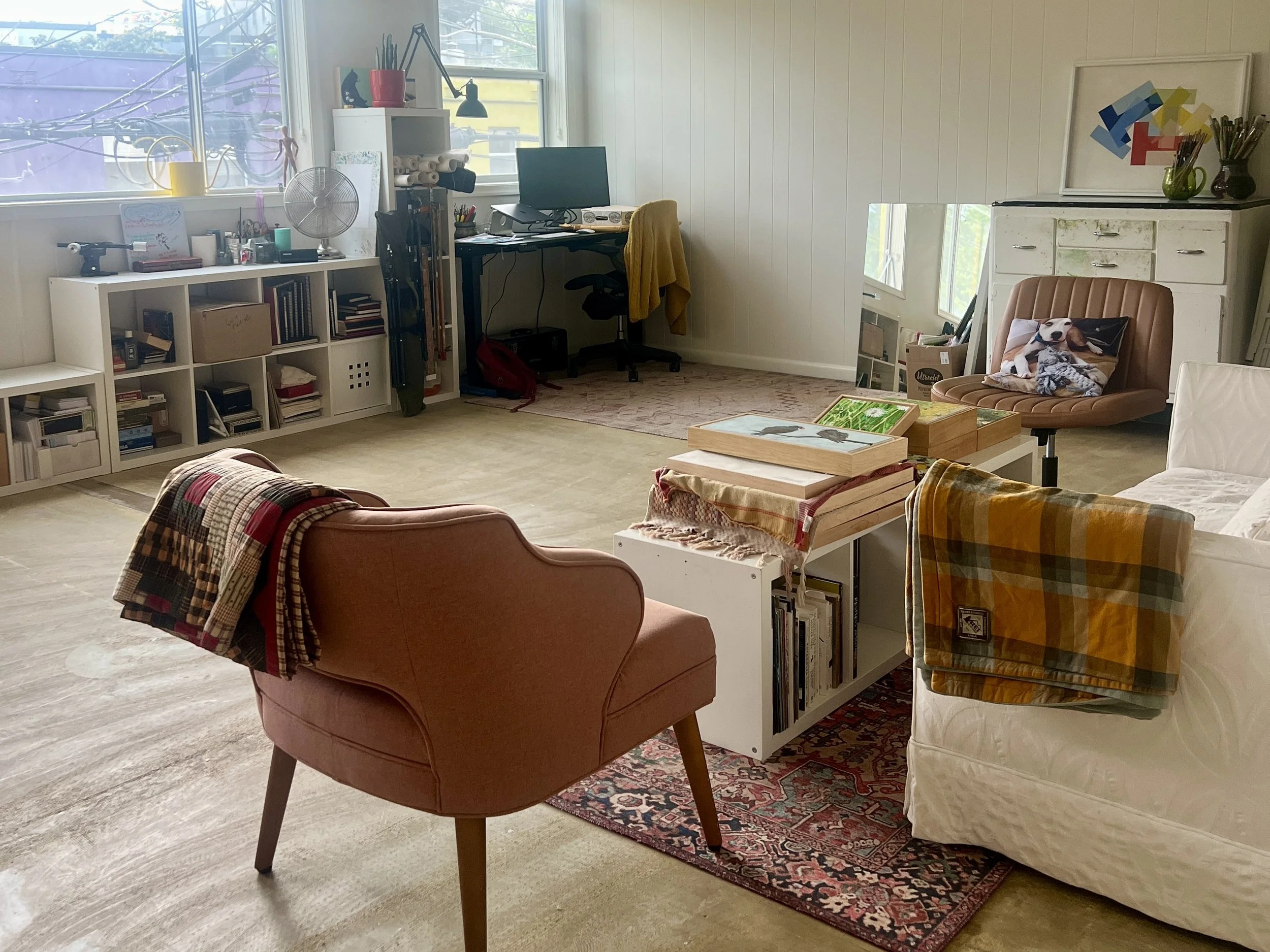 Art studio corner with pink armchair with patchwork blanket, white bookshelf, computer desk with monitor, framed art, mirror, and various decor items.
