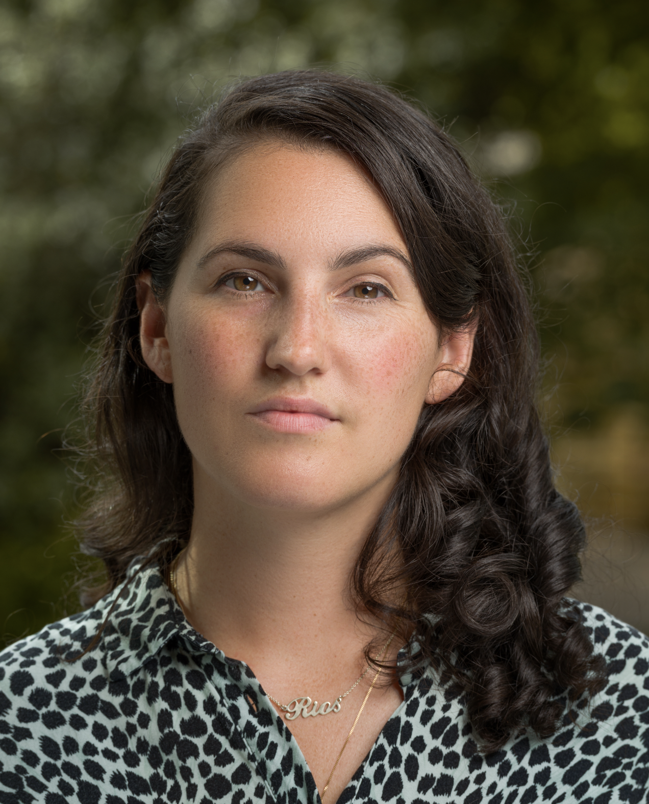 Close-up of a woman with shoulder-length wavy dark hair, wearing a leopard print blouse and a gold necklace with a name pendant that says 'Rius', outdoors with blurred green foliage in the background.
