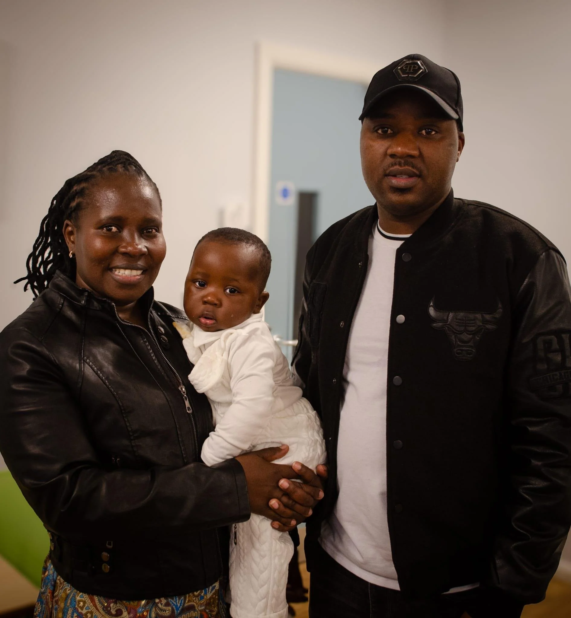 A woman holding a baby, standing next to two men in an indoor setting with white and green walls.