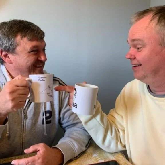 Two men smiling and clinking coffee mugs in a casual setting, engaged in conversation.