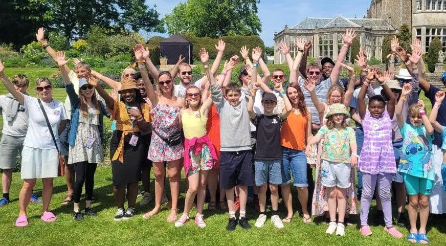 Group of children and adults outdoors on a sunny day with arms raised, smiling, in front of a historic mansion-like building.