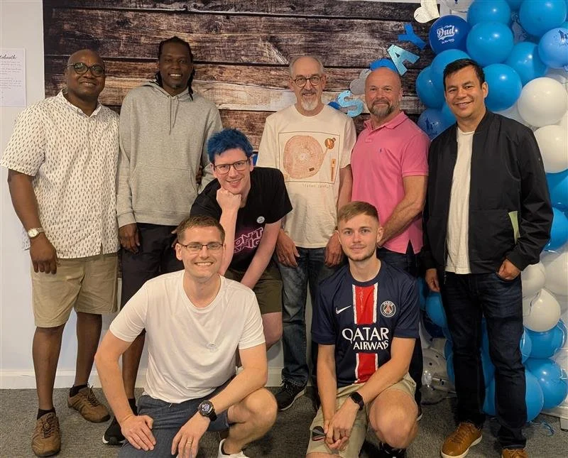 A group of nine diverse people posing together during an event, with balloons and a wooden backdrop in the background.