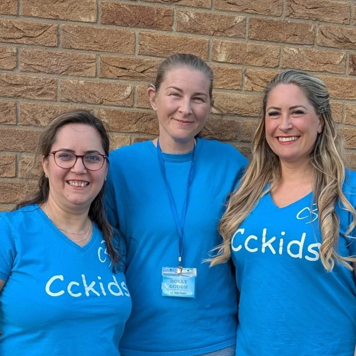 Three women standing arm-in-arm in front of a brick wall, smiling at the camera
