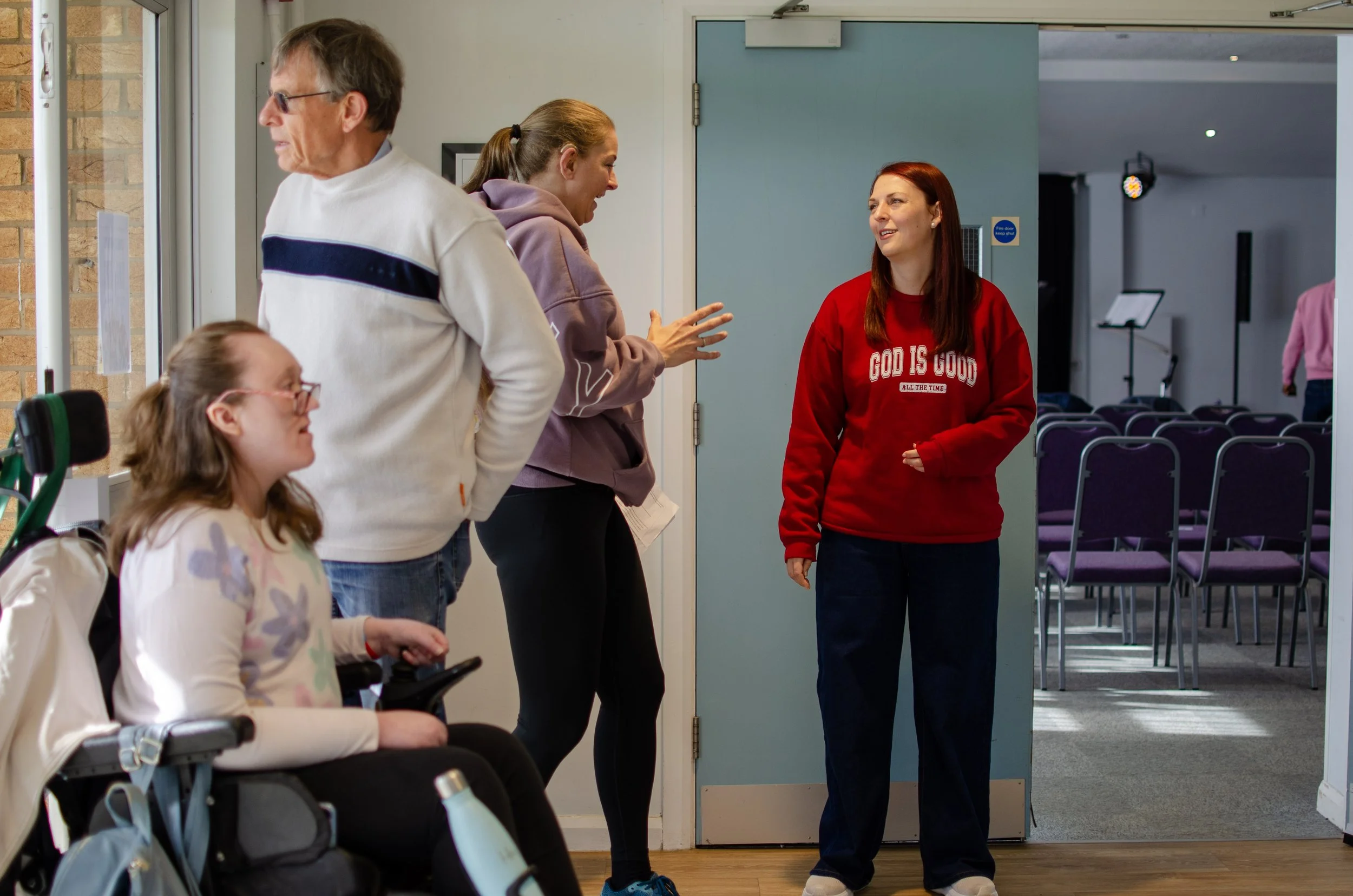 A group of people, including a young girl in a wheelchair, are interacting with two women in a building lobby. One woman is wearing a purple hoodie and is gesturing as she talks, while the other woman is smiling and wearing a red sweatshirt that says "GOD IS GOOD ALL THE TIME." There are purple chairs arranged in rows in the background and a person in a pink jacket walking away.
