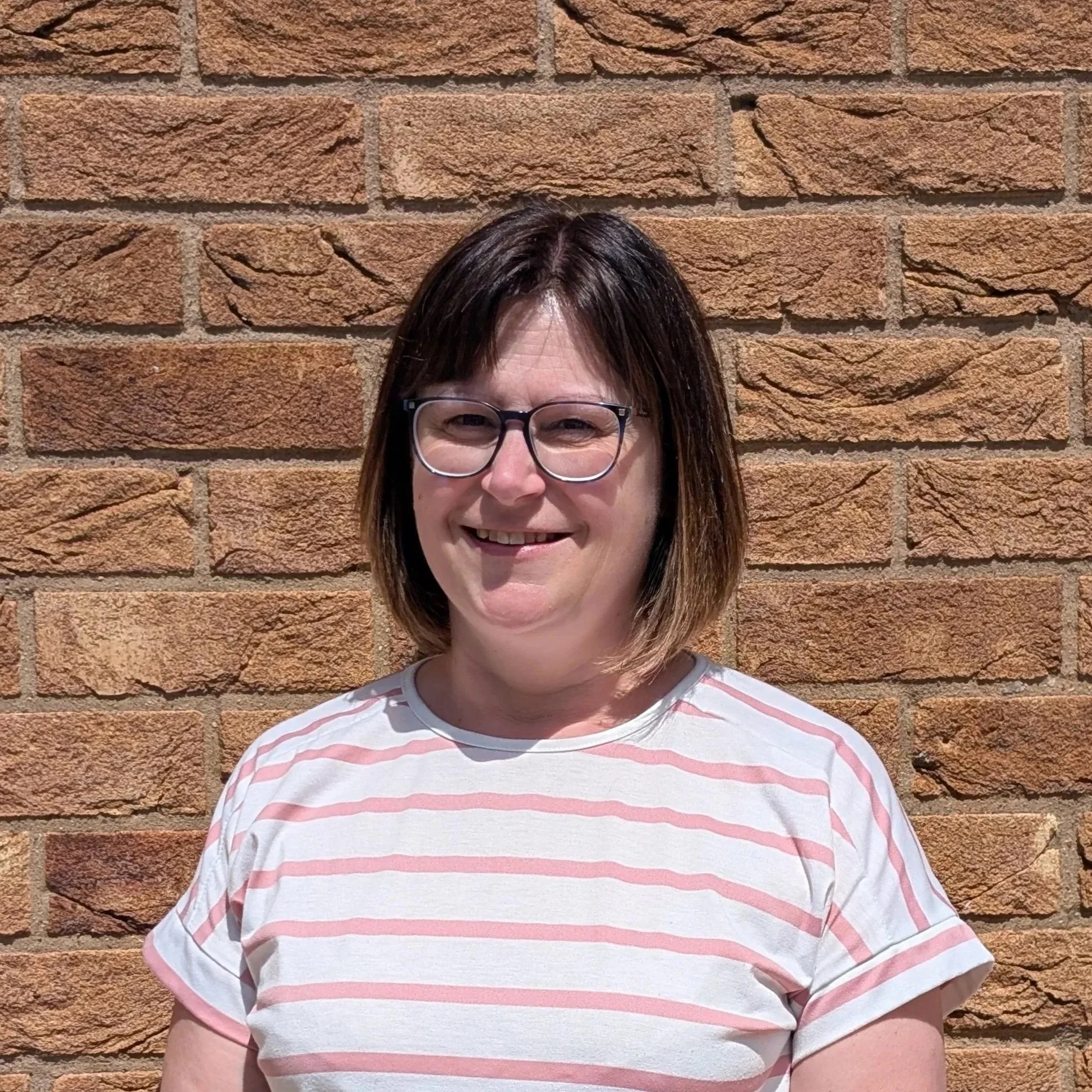 A lady with glasses and brown hair smiling in front of a brick wall