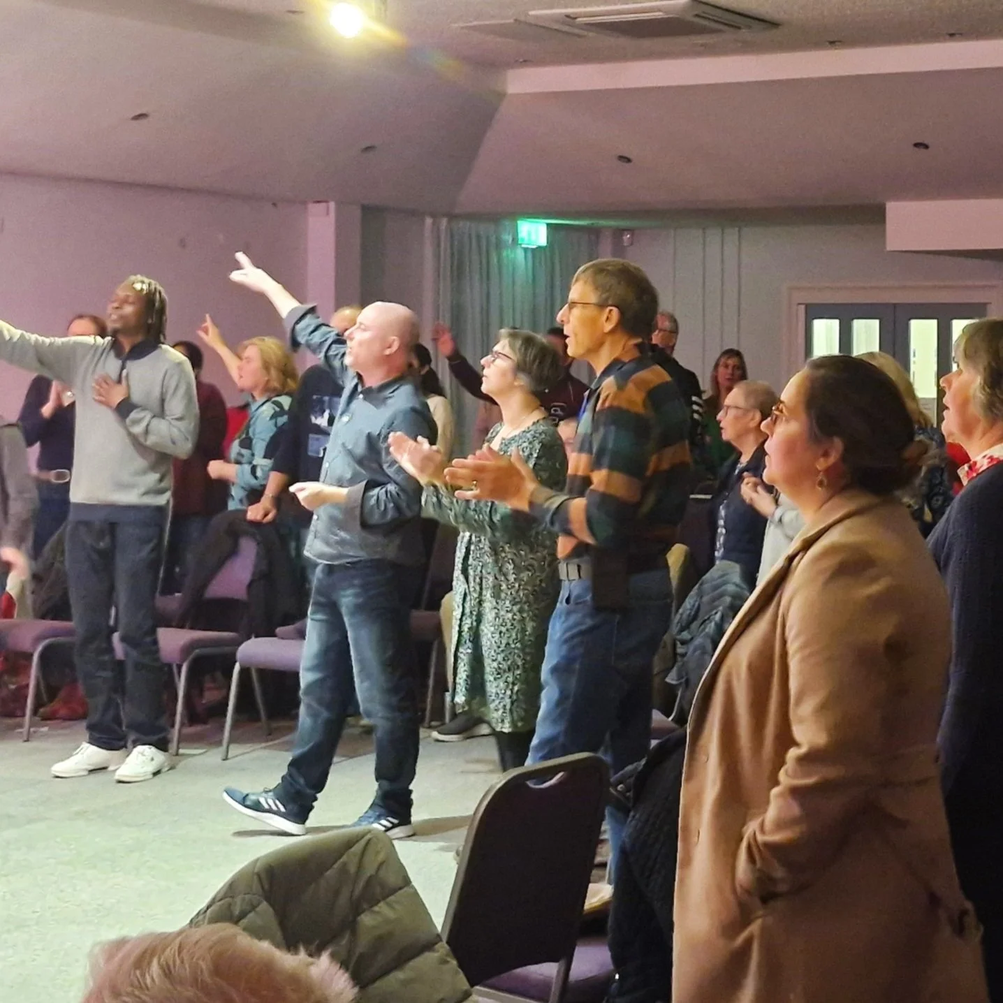 A group of people standing and clapping in a room, likely at a church or conference, with some raising their hands and others singing or praying.