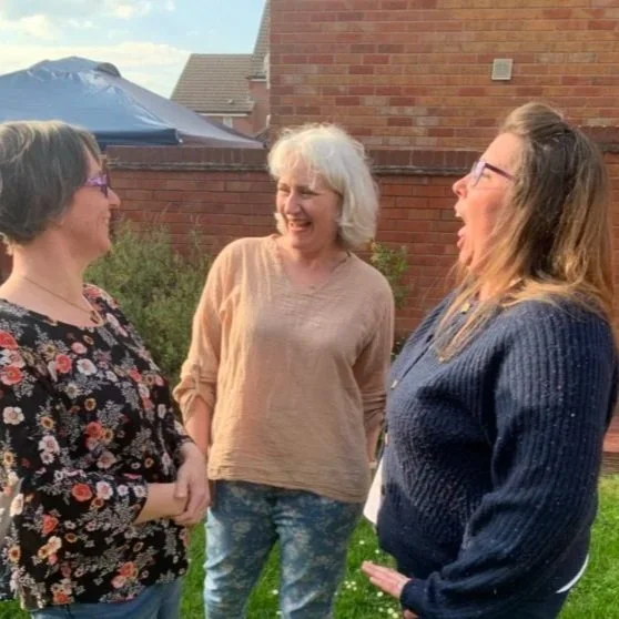 Three women with glasses and long hair standing and talking outside on a grassy lawn, smiling and laughing near a brick house with a blue tent in the background.