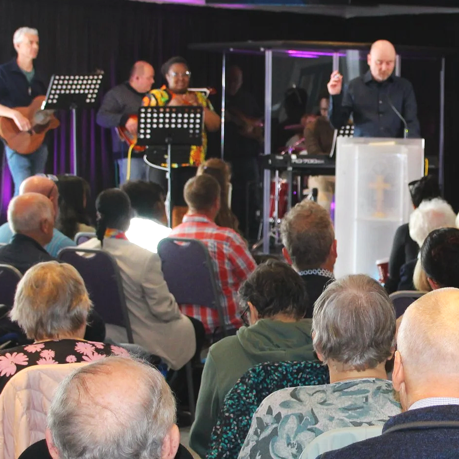 A group of people sitting in chairs, attending a church service or concert, with several musicians on stage playing instruments.