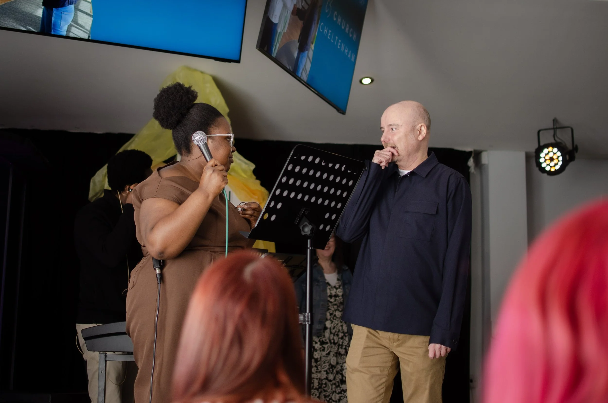 A woman with braided hair holding a microphone and speaking or singing on stage alongside a bald man. 