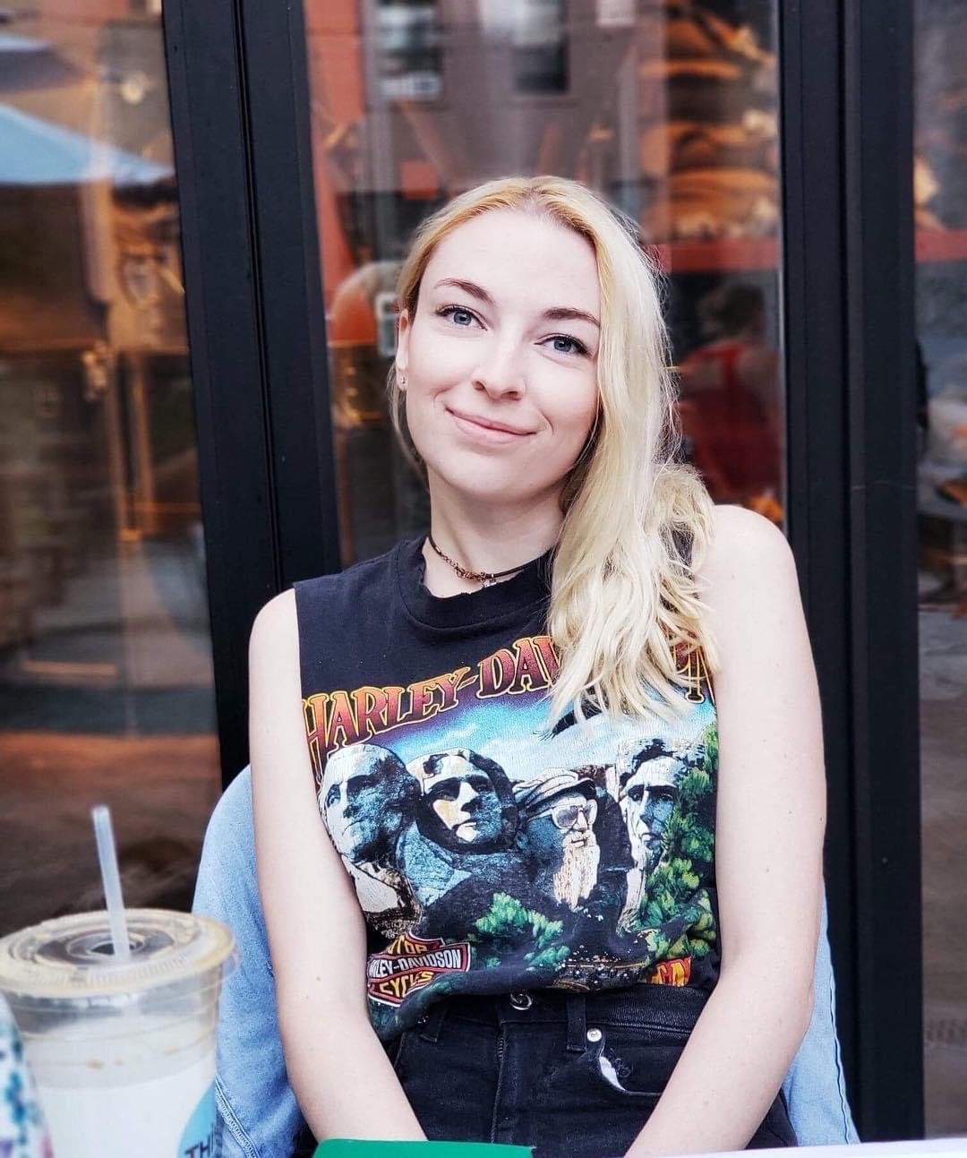 A young woman with long blonde hair smiling, wearing a sleeveless Harley-Davidson t-shirt, sitting outdoors near a glass window or door.