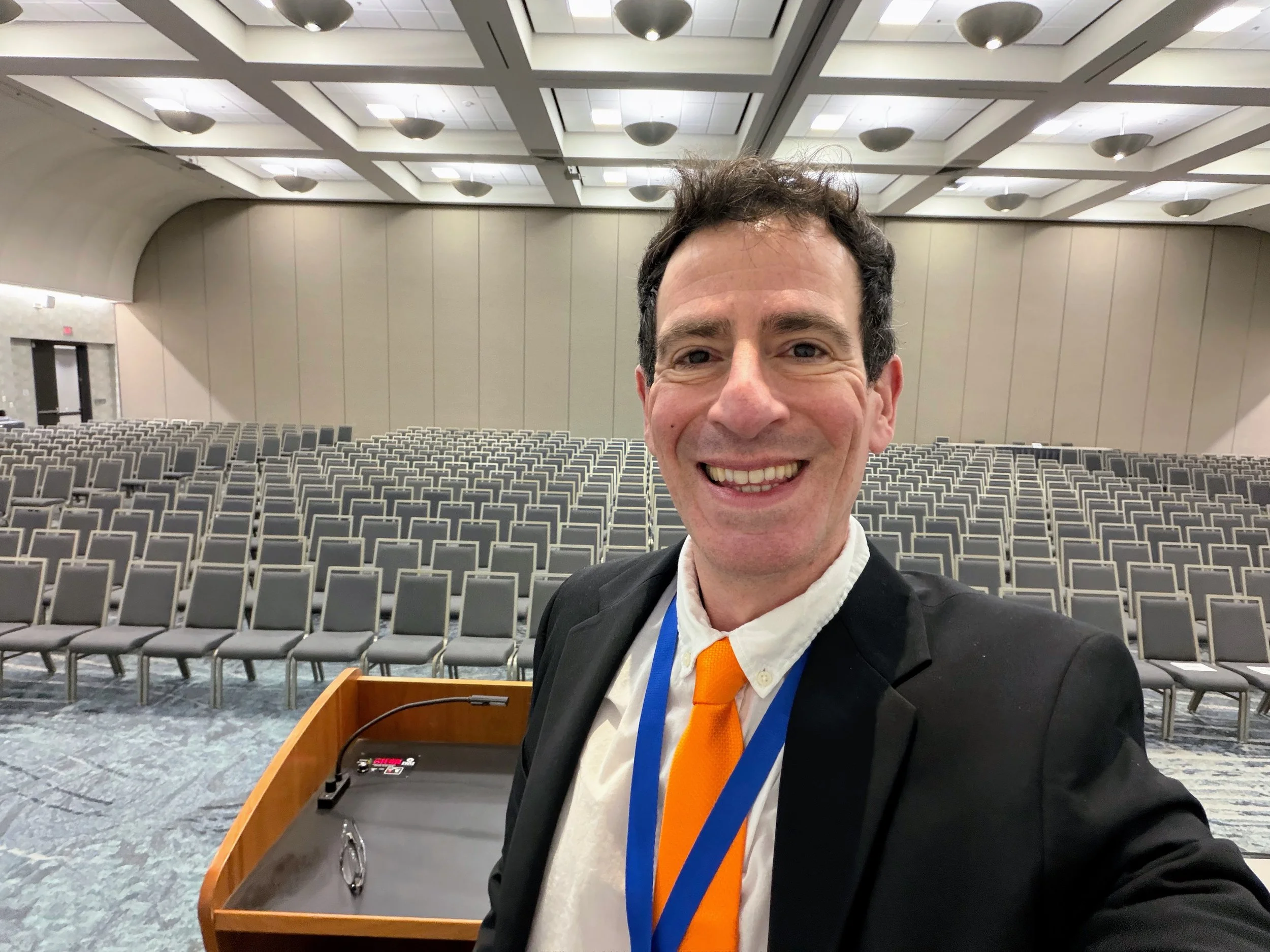Dr. Gregory Charlop, seen here in a large conference room wearing a black sport coat and orange tie,  was a featured speaker at the WEST 2026 Naval Warfare conference