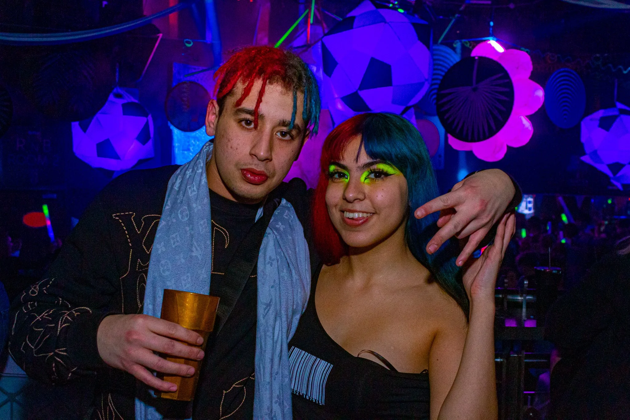 Two people with colorful hair and neon makeup in a nightclub with geometric decorations.