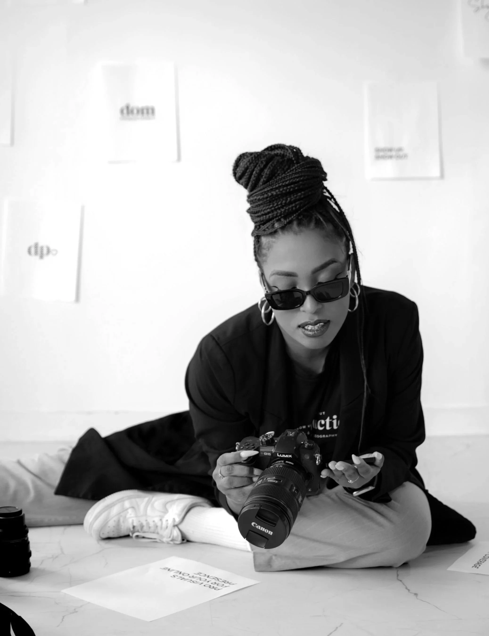 Black and white photo of a woman with braided hair in a bun, wearing sunglasses, hoop earrings, a dark jacket, white sneakers, and gray pants, sitting on the floor holding a camera and looking at her phone, with a camera lens on the floor nearby.