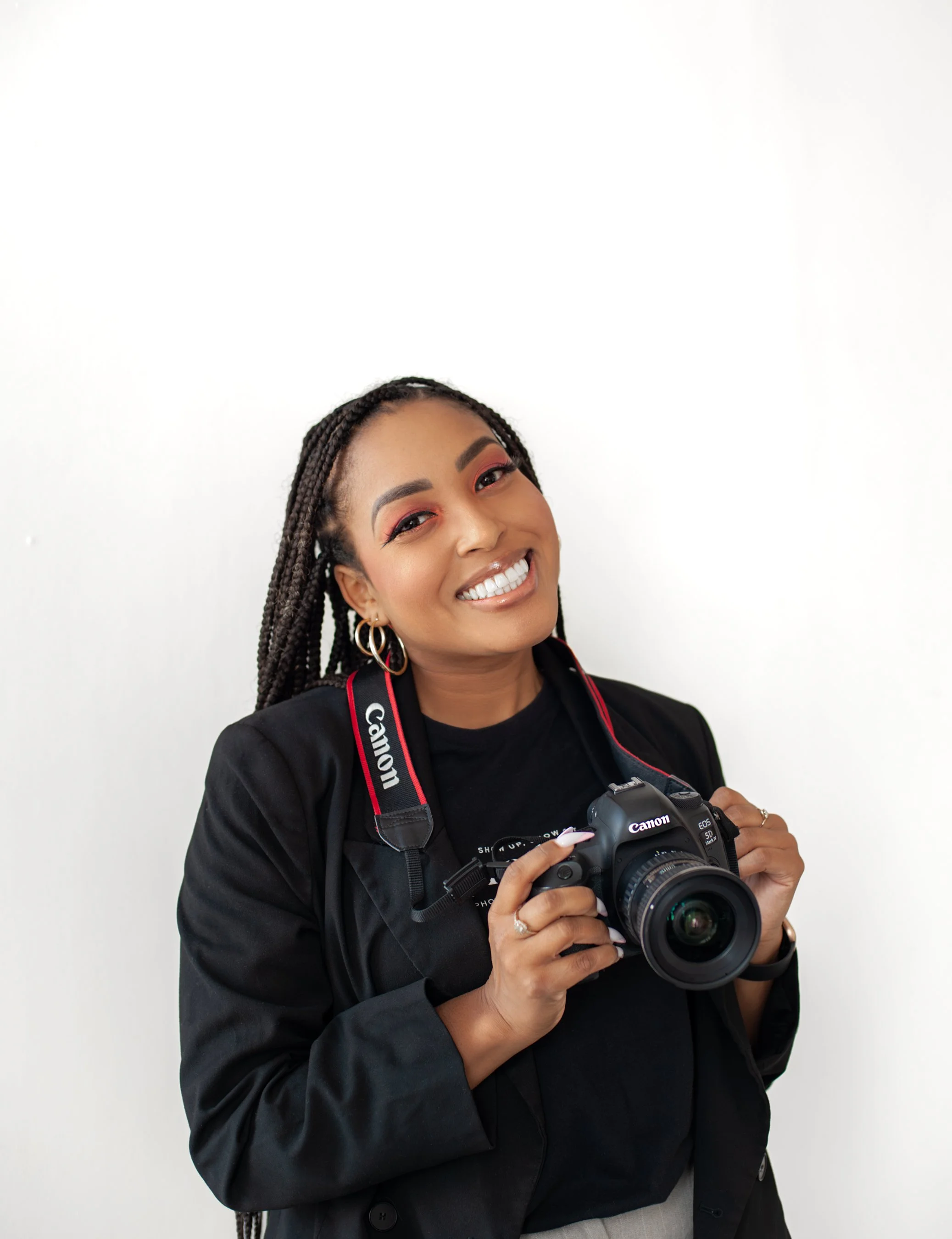 A woman with curly hair, wearing a black blazer and leather pants, sitting on a chair in a photography studio, smiling at the camera with photography equipment around her.