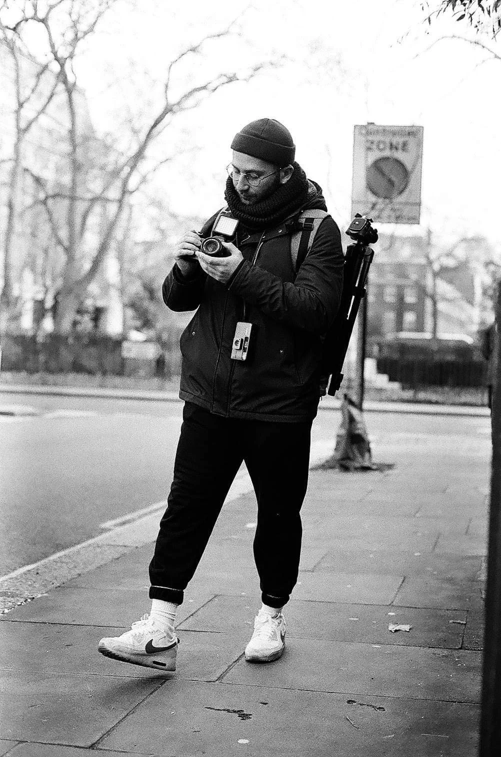 A young man in casual clothing, including a beanie, glasses, and sneakers, standing on a sidewalk and looking at his camera. He has a backpack with a tripod attached and a lanyard with a badge around his neck. The scene is in black and white, with trees and a street sign in the background.
